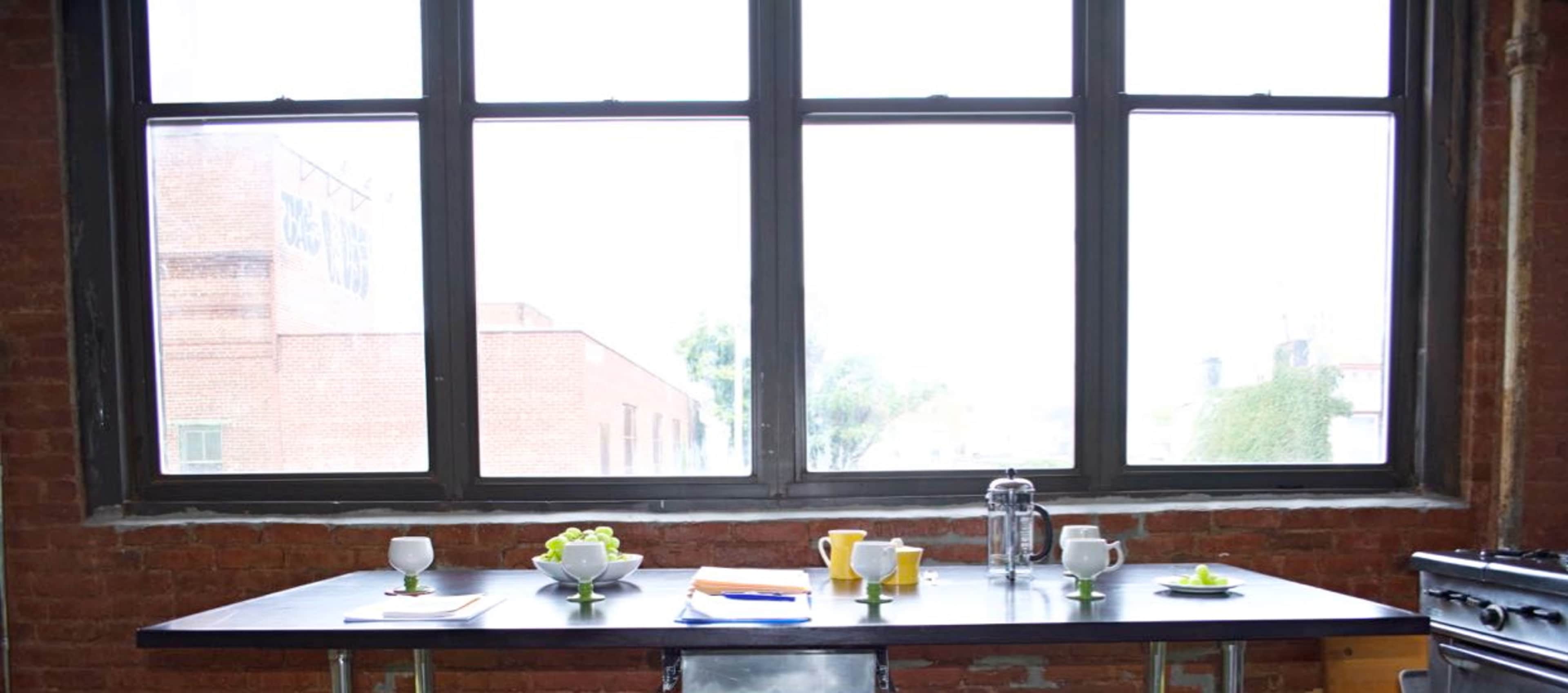 A wooden table is set with dishes and a coffee press, positioned in front of large windows in a brick-walled room.