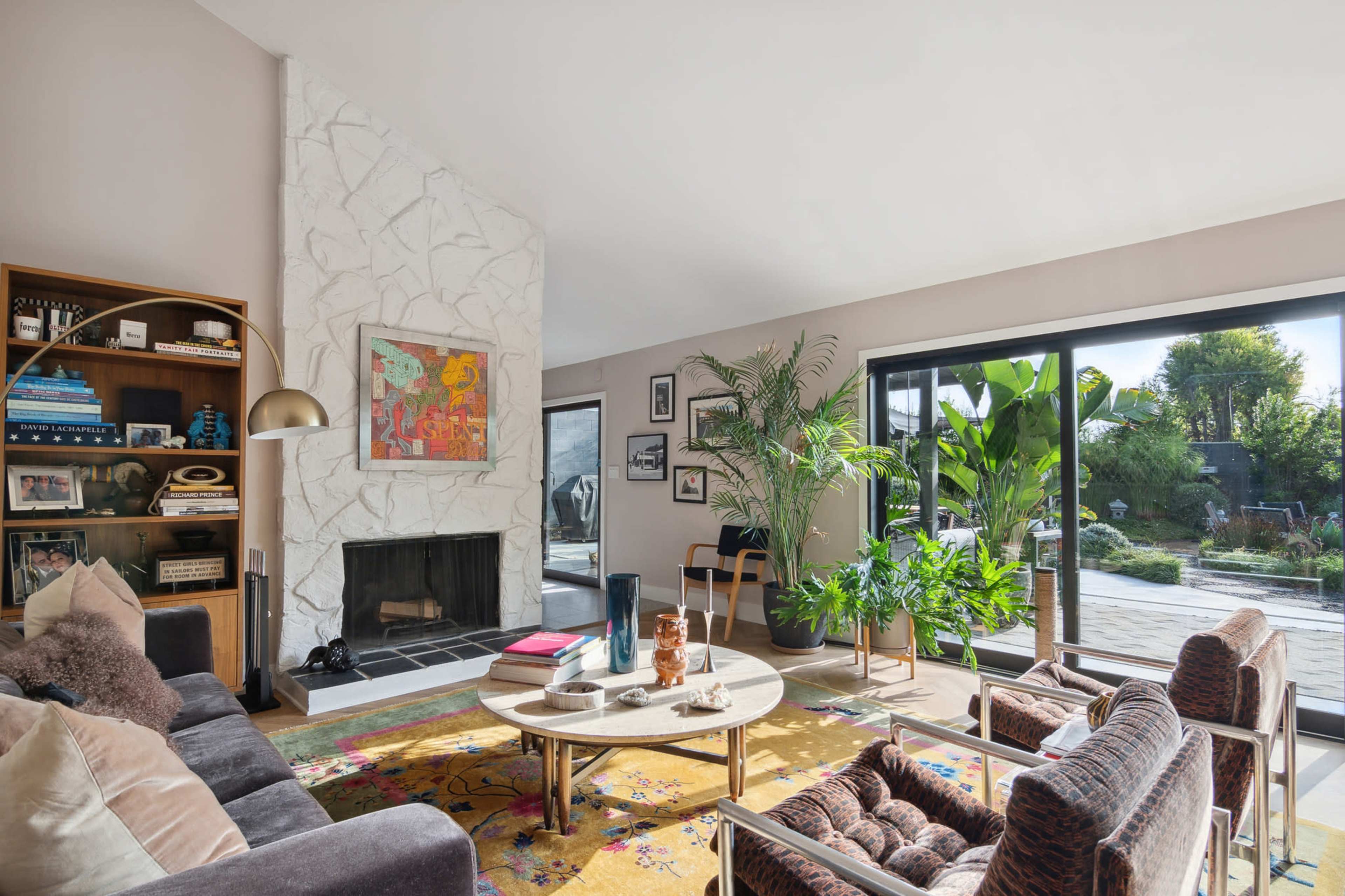 A modern living room features a stone fireplace, a colorful area rug, and large windows that open to a garden.
