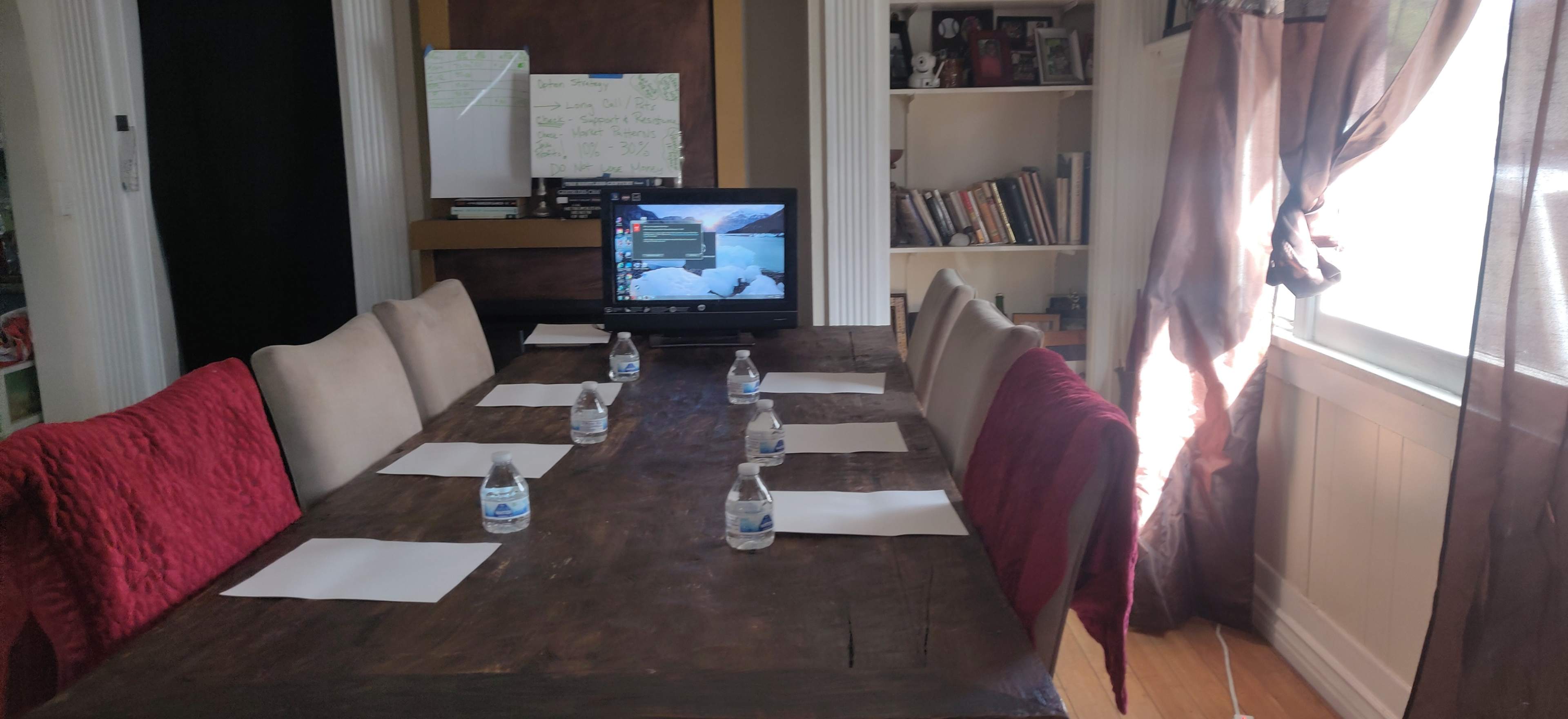 A long wooden dining table is set with bottles of water and blank papers, while a television displaying a menu screen is positioned at one end of the room.