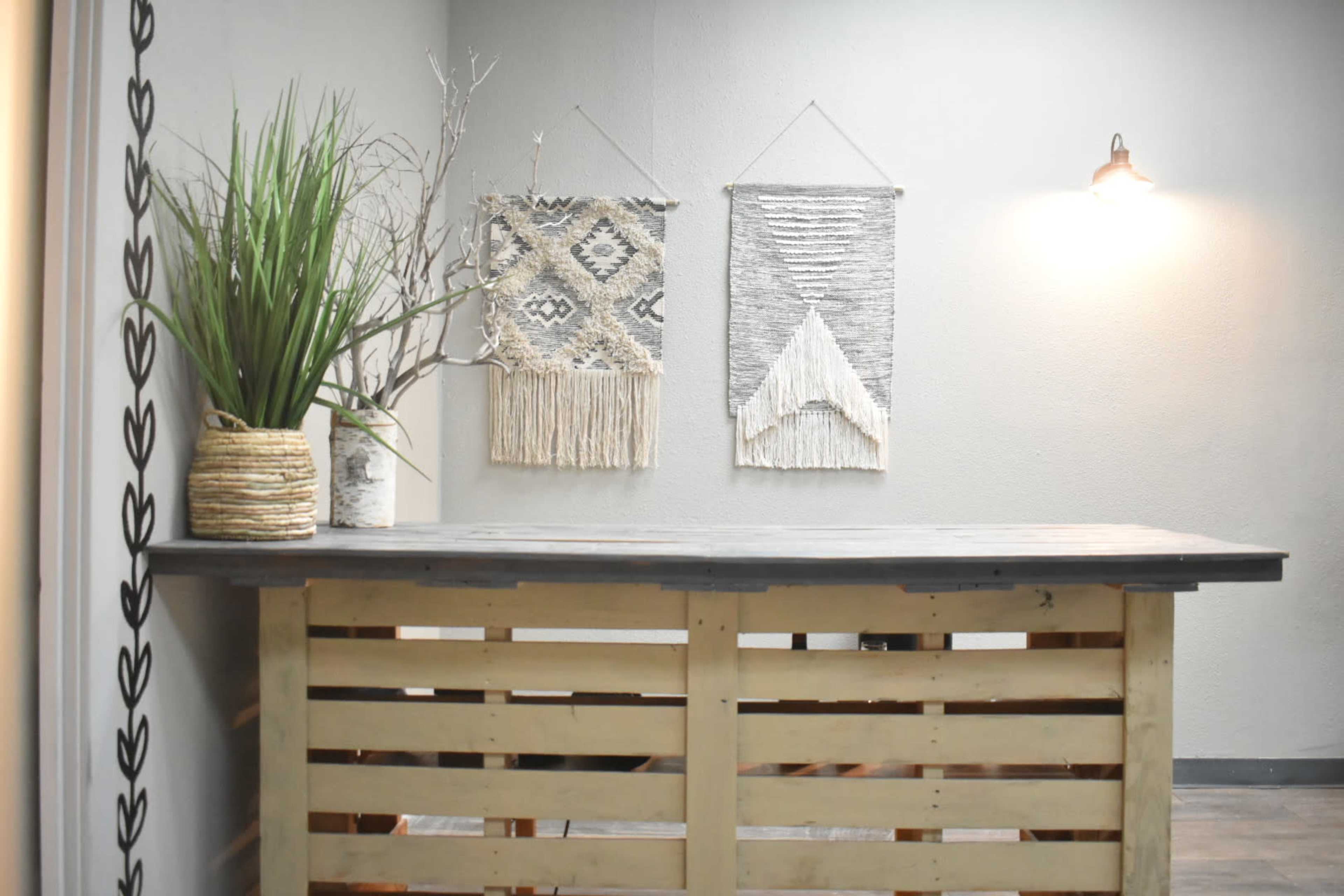 A wooden counter made of pallet wood in front of a wall adorned with two woven wall hangings and a potted plant.