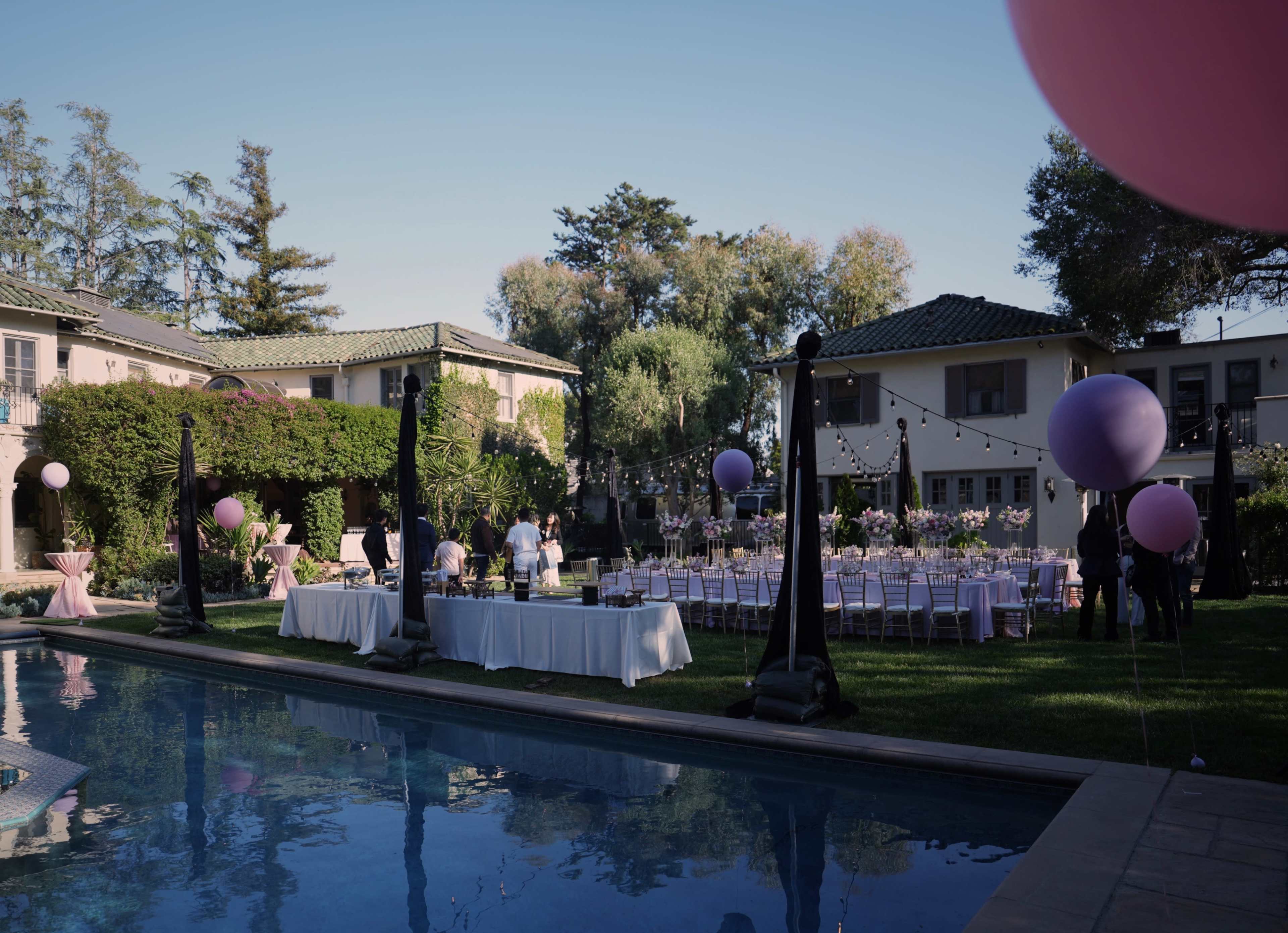 The scene depicts an outdoor event setup with tables adorned for a gathering beside a swimming pool, surrounded by trees and elegant decorations.