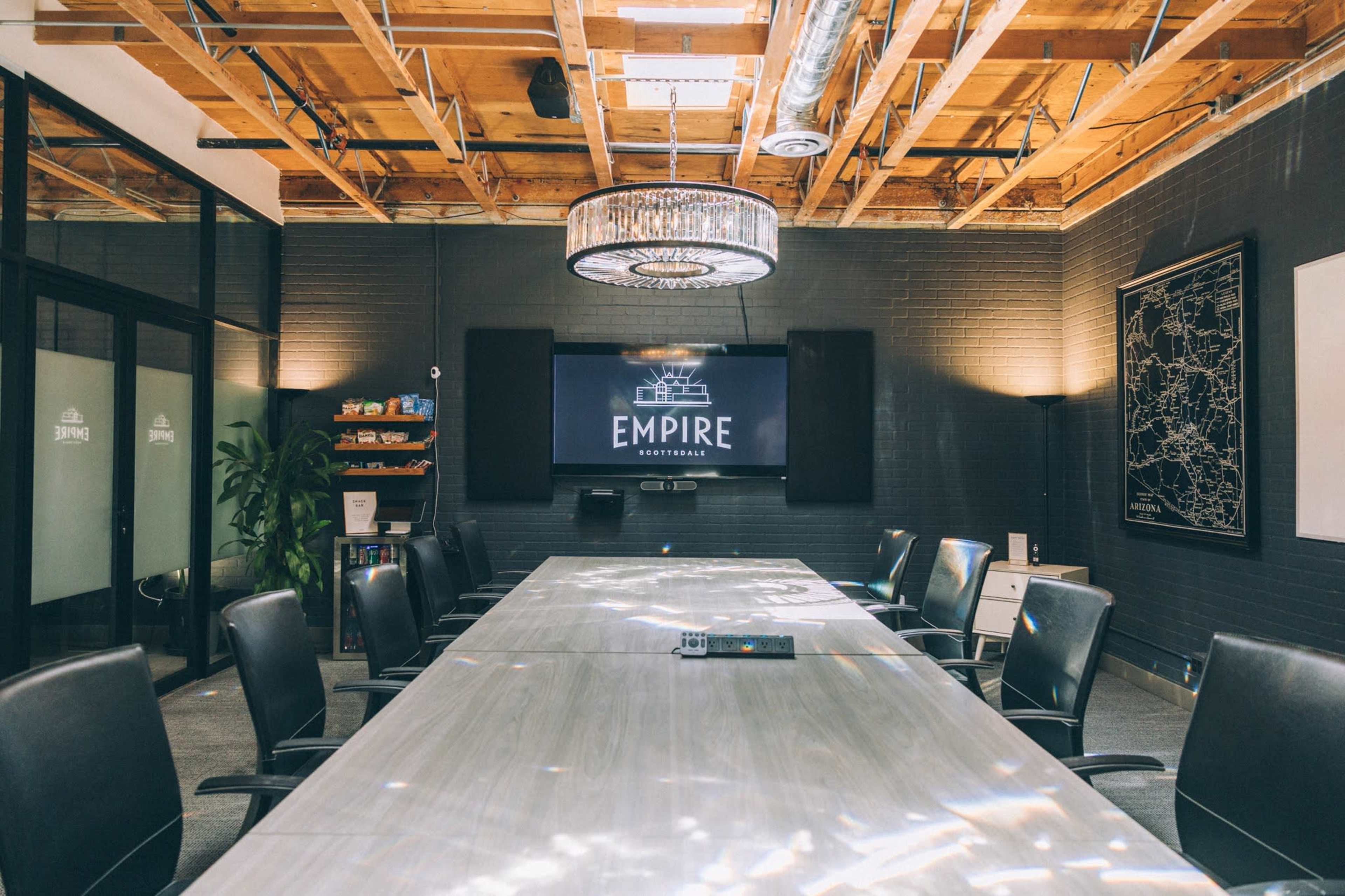 A modern conference room features a long table surrounded by black chairs, a large screen displaying the word "EMPIRE," and exposed wooden beams overhead.