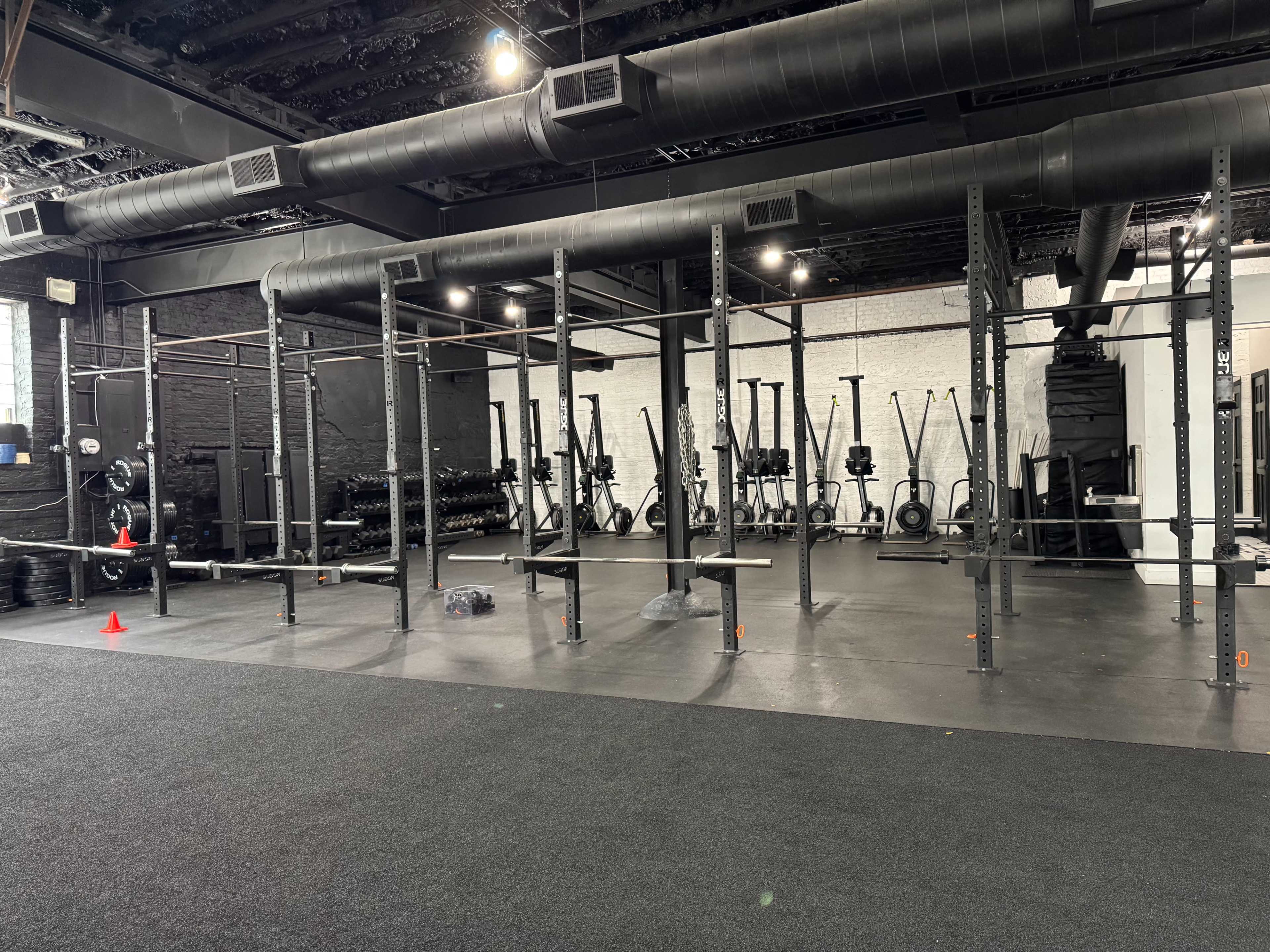 The image shows a spacious gym interior featuring multiple exercise racks, rowing machines, and dumbbells against a black and white backdrop.