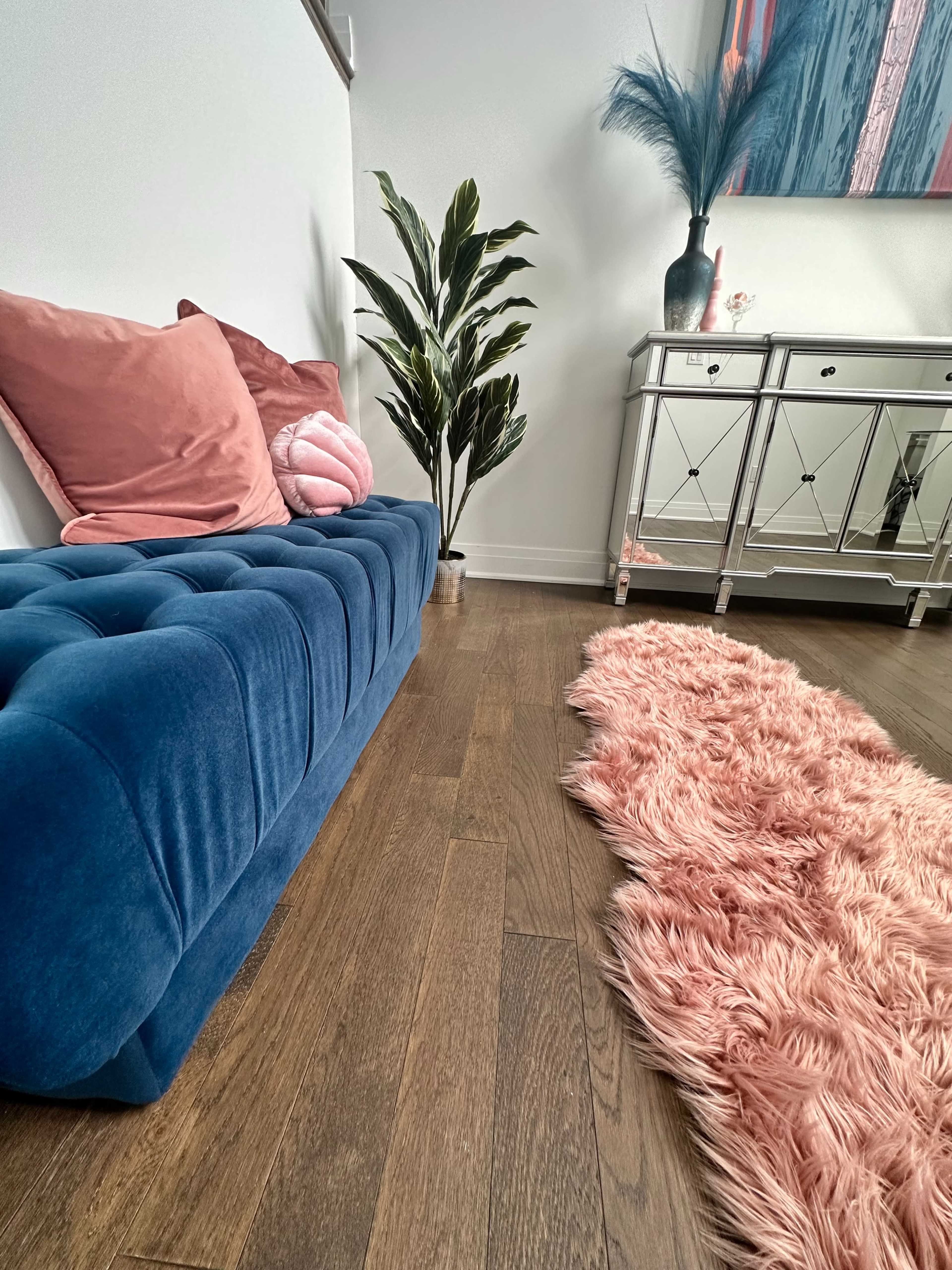 A blue tufted sofa is positioned next to a decorative plant and a mirrored console table on a hardwood floor, accompanied by a pink shag rug.