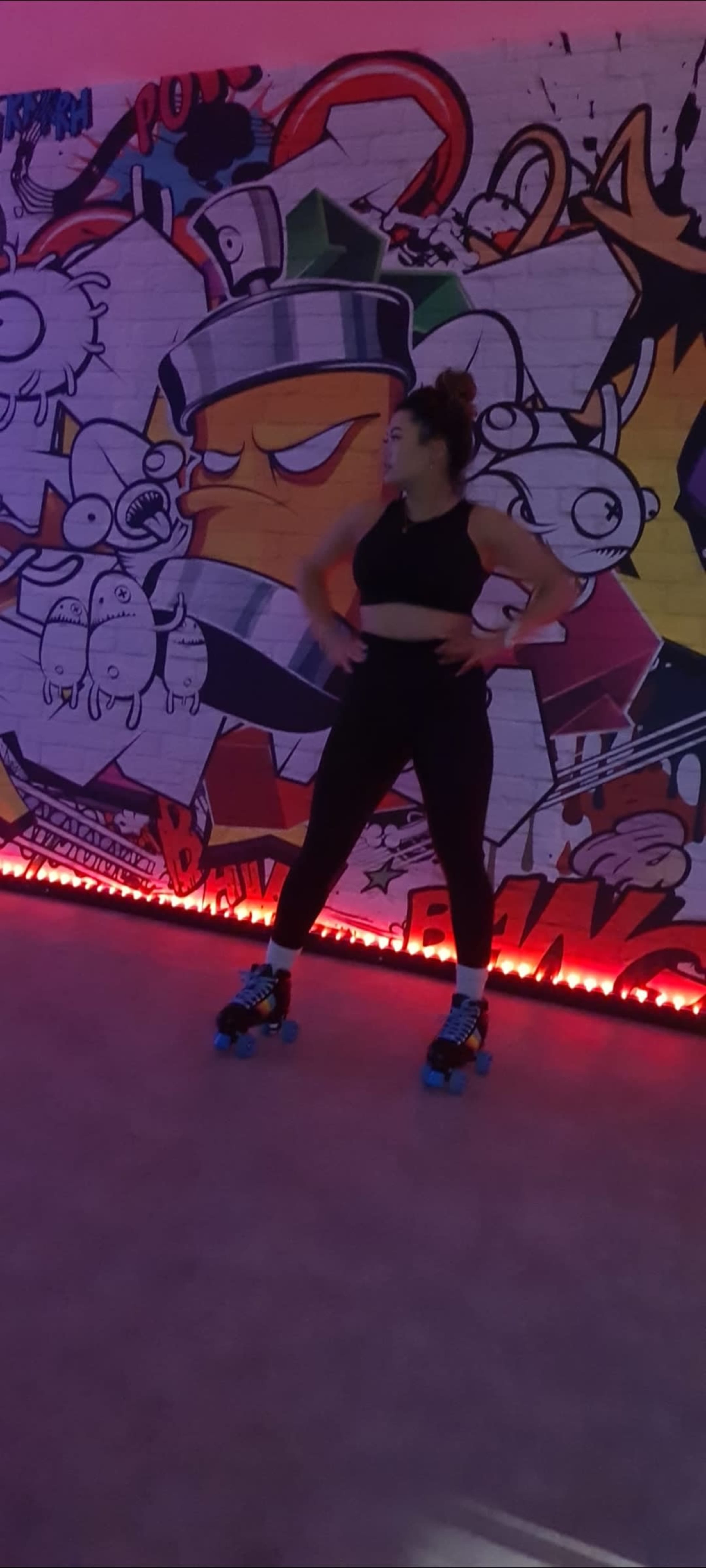 A woman wearing a crop top and black leggings performs on roller skates against a vibrant graffiti mural with colorful cartoon characters.