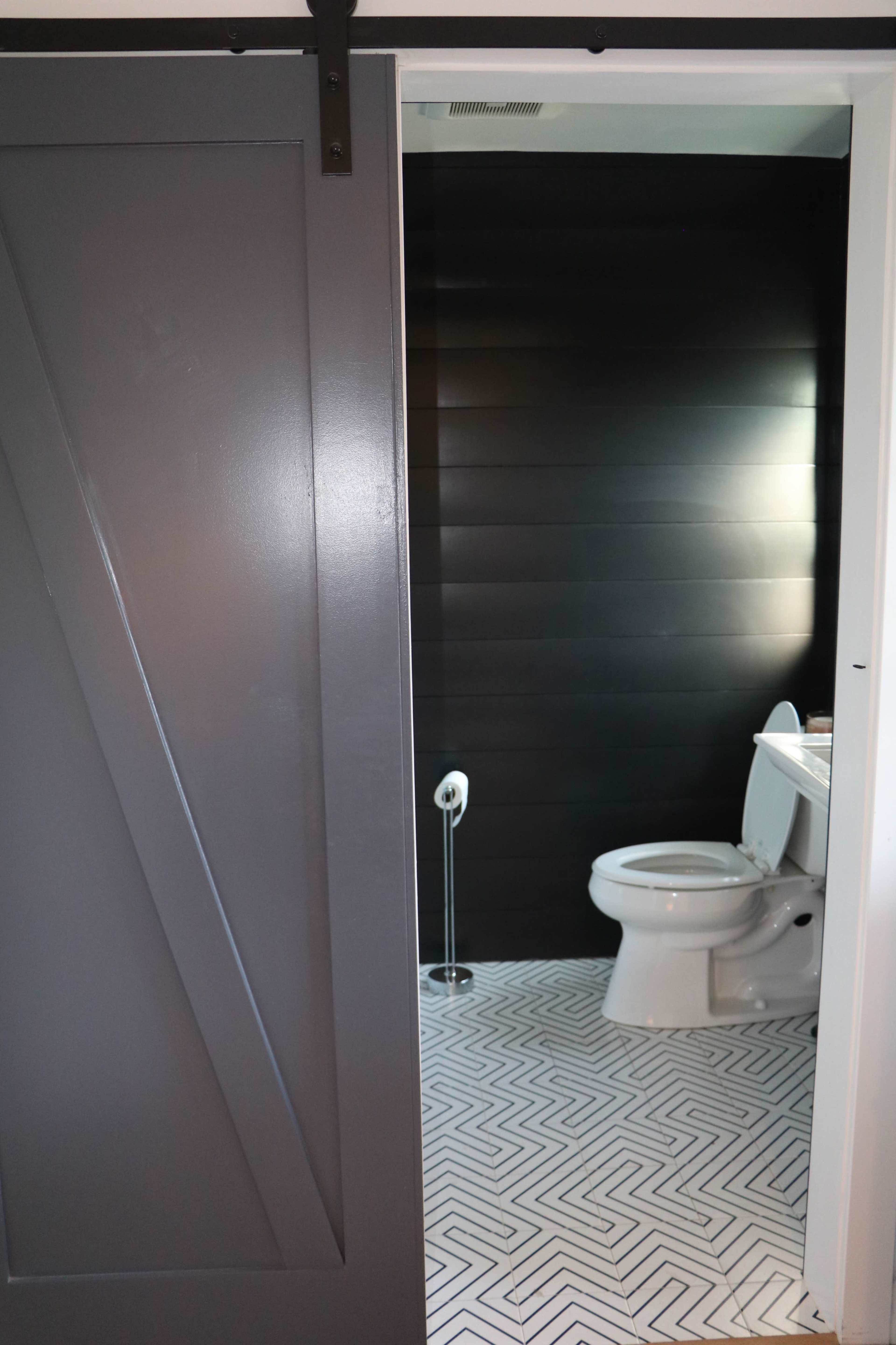 The image shows a modern bathroom with a sliding barn door, a black accent wall, and a white toilet on a patterned tile floor.