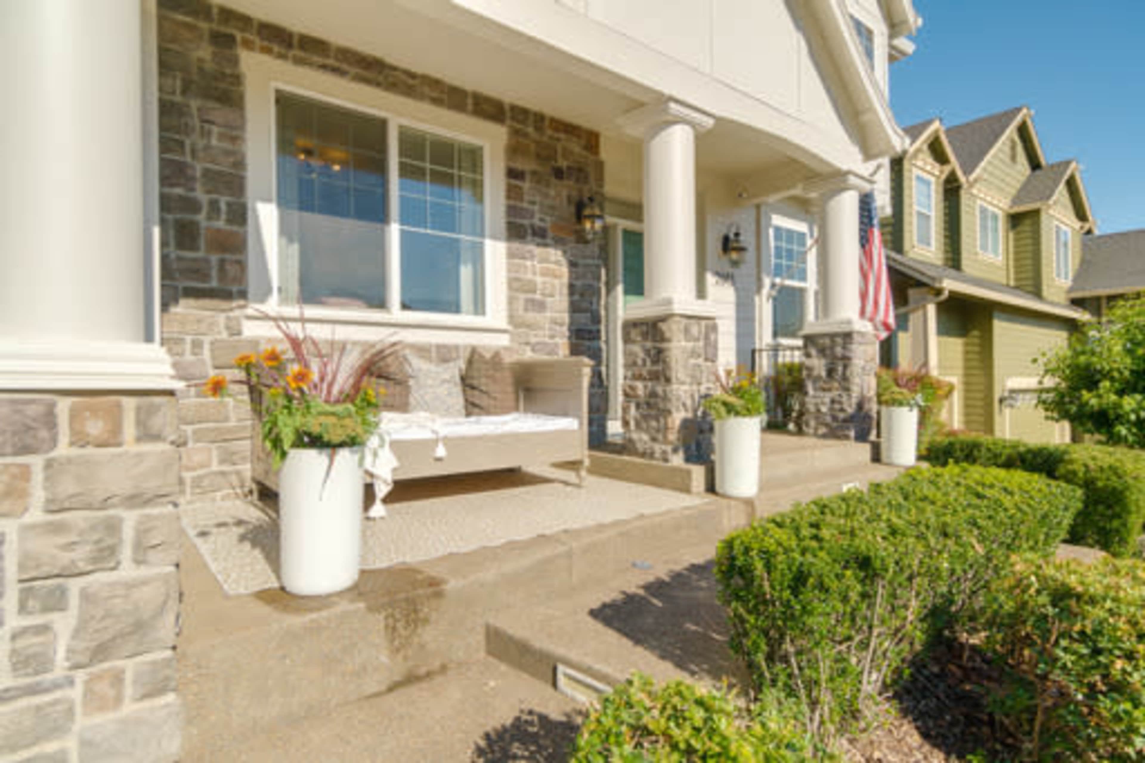 The front porch of a house features a stone facade, a bench, potted plants, and an American flag.