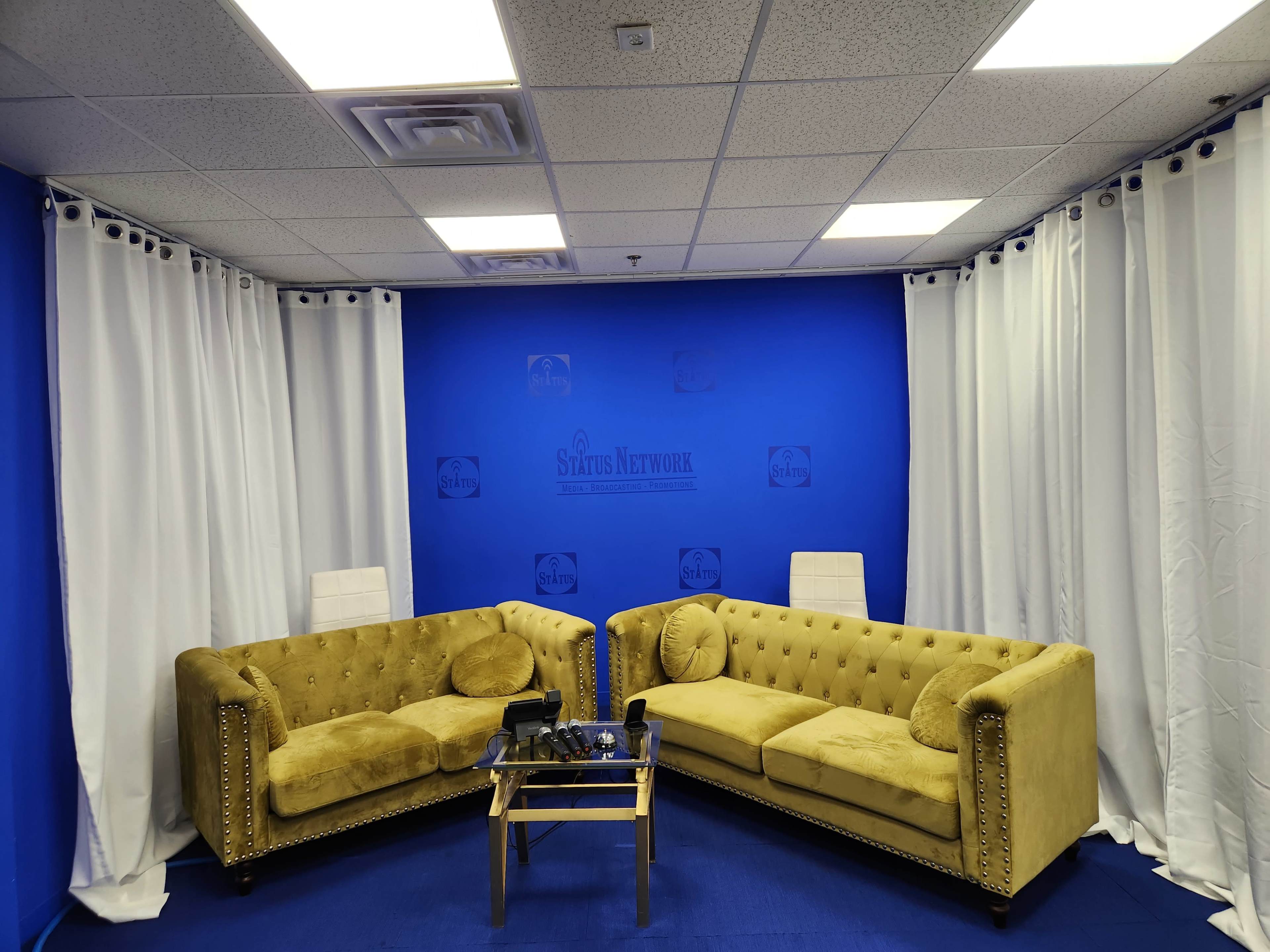 A studio-like setting with two yellow tufted sofas facing each other, a glass coffee table in the middle, and blue walls with white curtains.