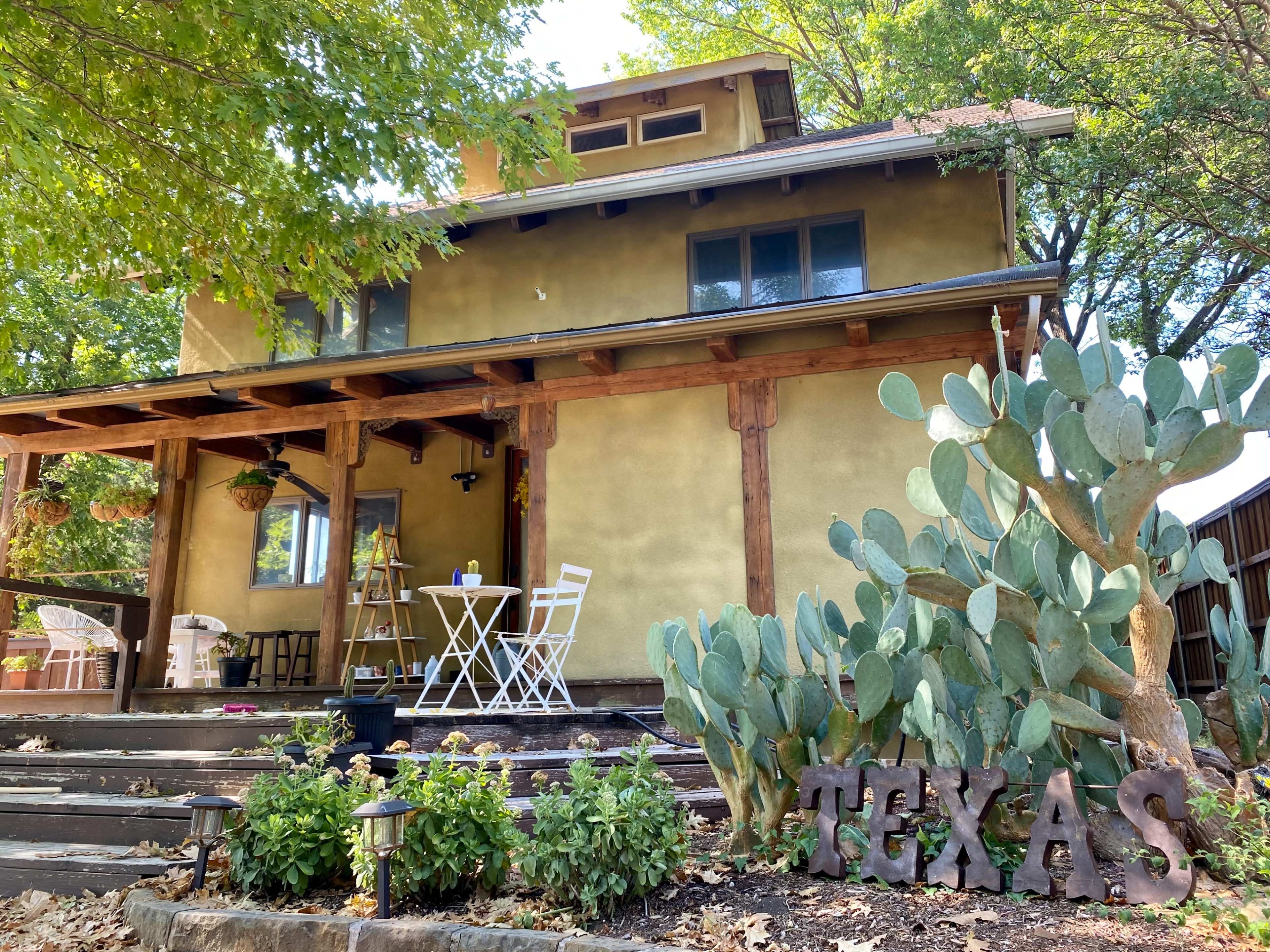 A two-story house with a greenish-yellow exterior is surrounded by trees and features a small outdoor seating area with a table and chairs, alongside a cactus garden and a wooden sign spelling "TEXAS."