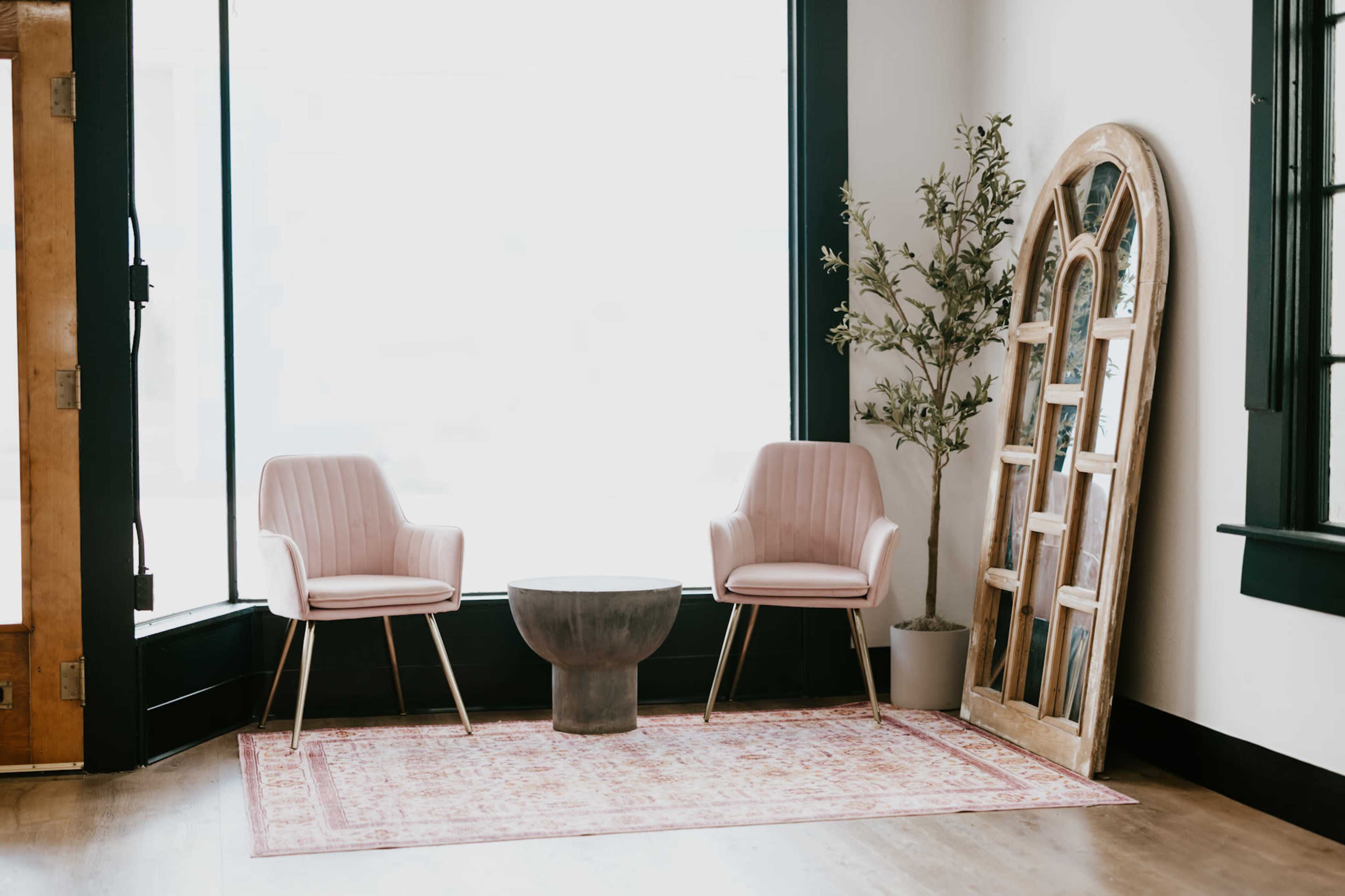 The image shows a minimalist interior with two pink chairs, a round table, a potted plant, and a large decorative mirror against a bright window.