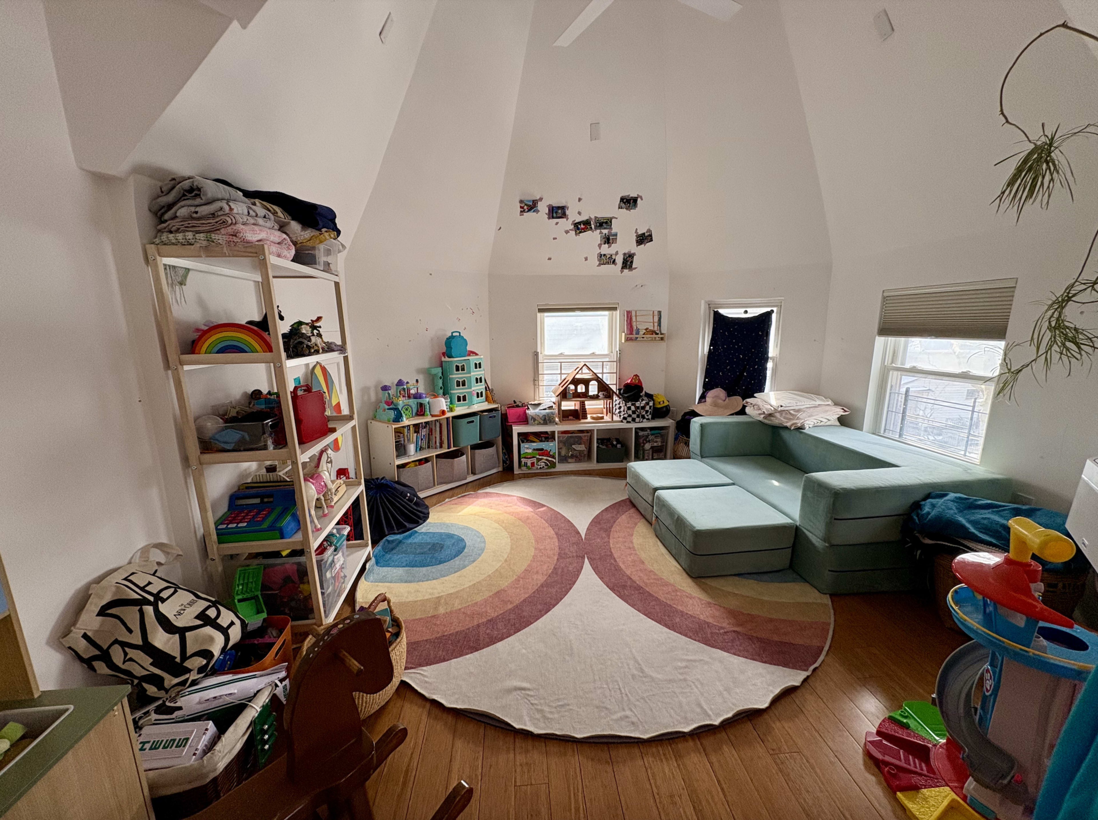 The image shows a brightly lit playroom with a colorful circular rug, shelves filled with toys, and a set of green couches arranged in an inviting way.