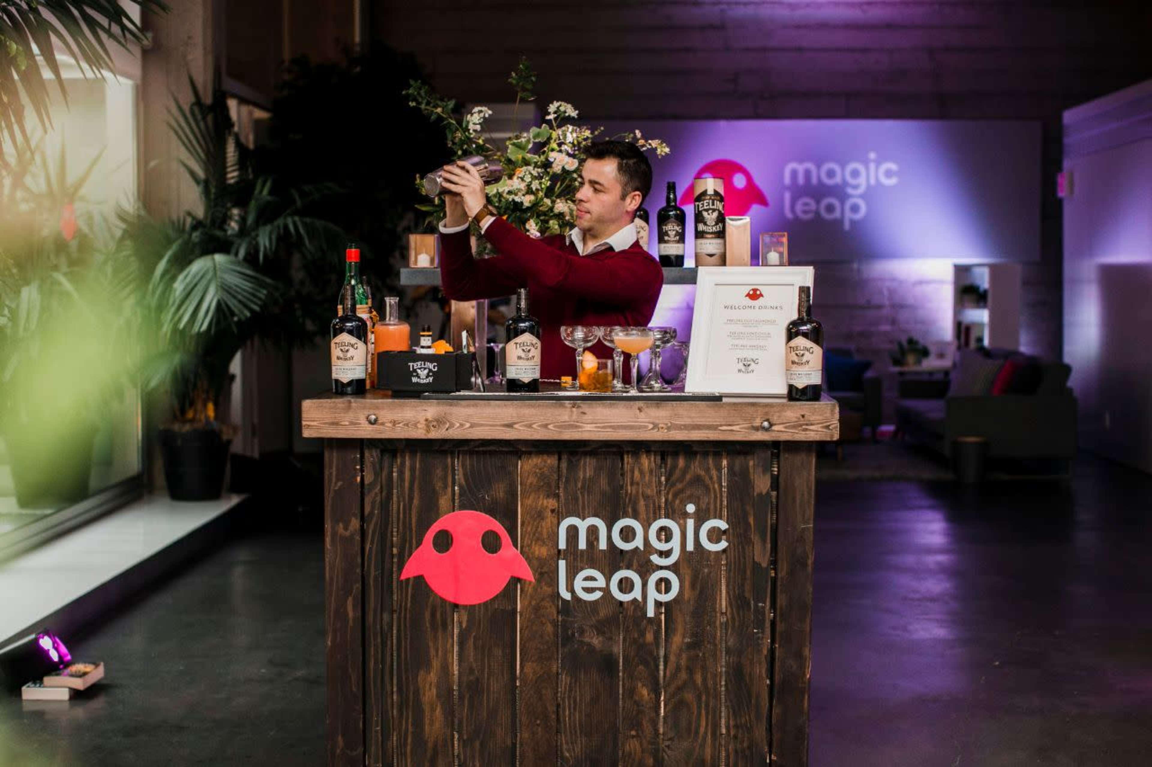A bartender prepares drinks at a sleek wooden bar featuring the "magic leap" logo and surrounded by plants in a modern setting.