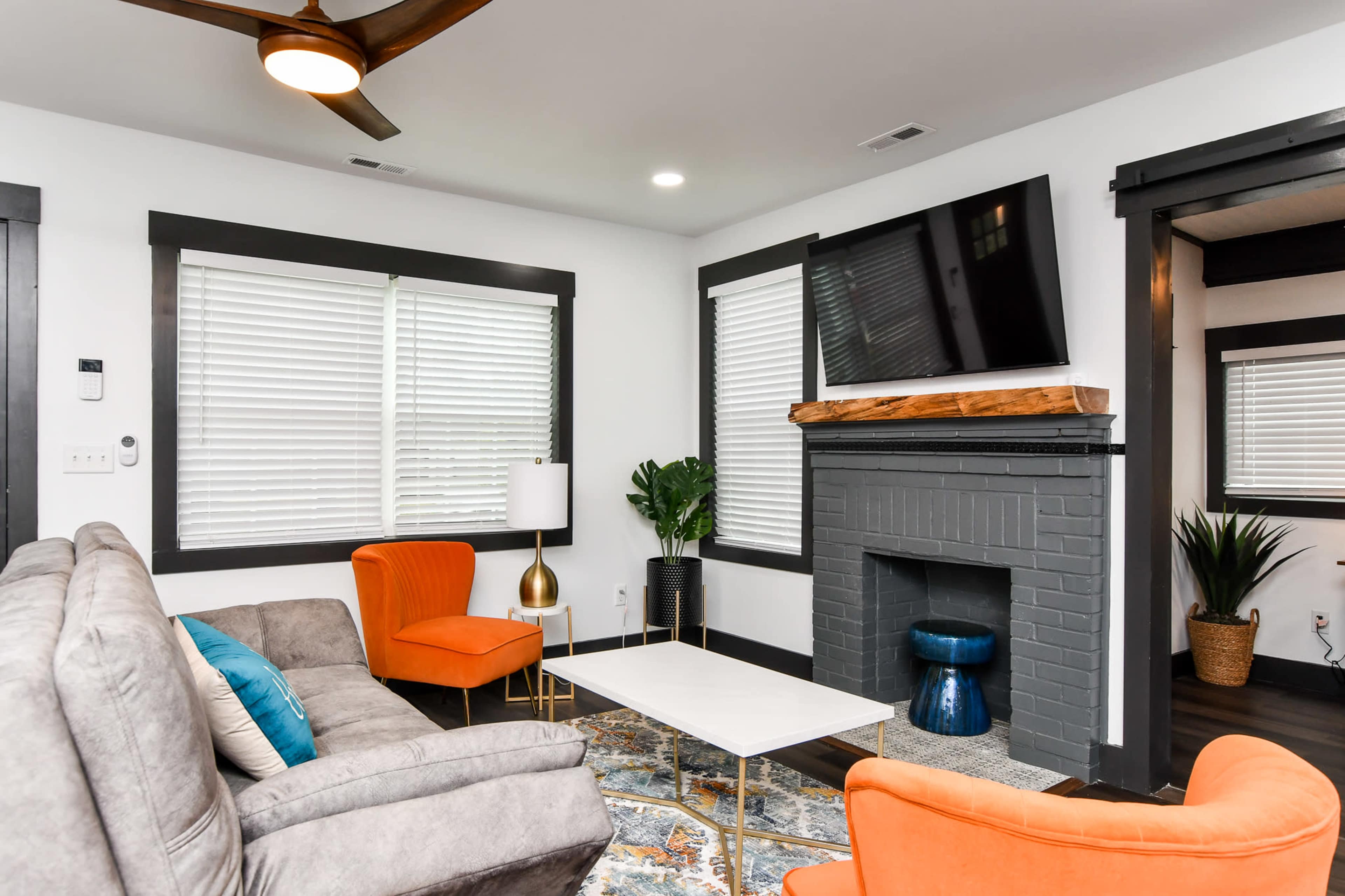 A modern living room featuring a gray sofa, two orange chairs, a coffee table, and a flat-screen TV mounted above a gray fireplace.