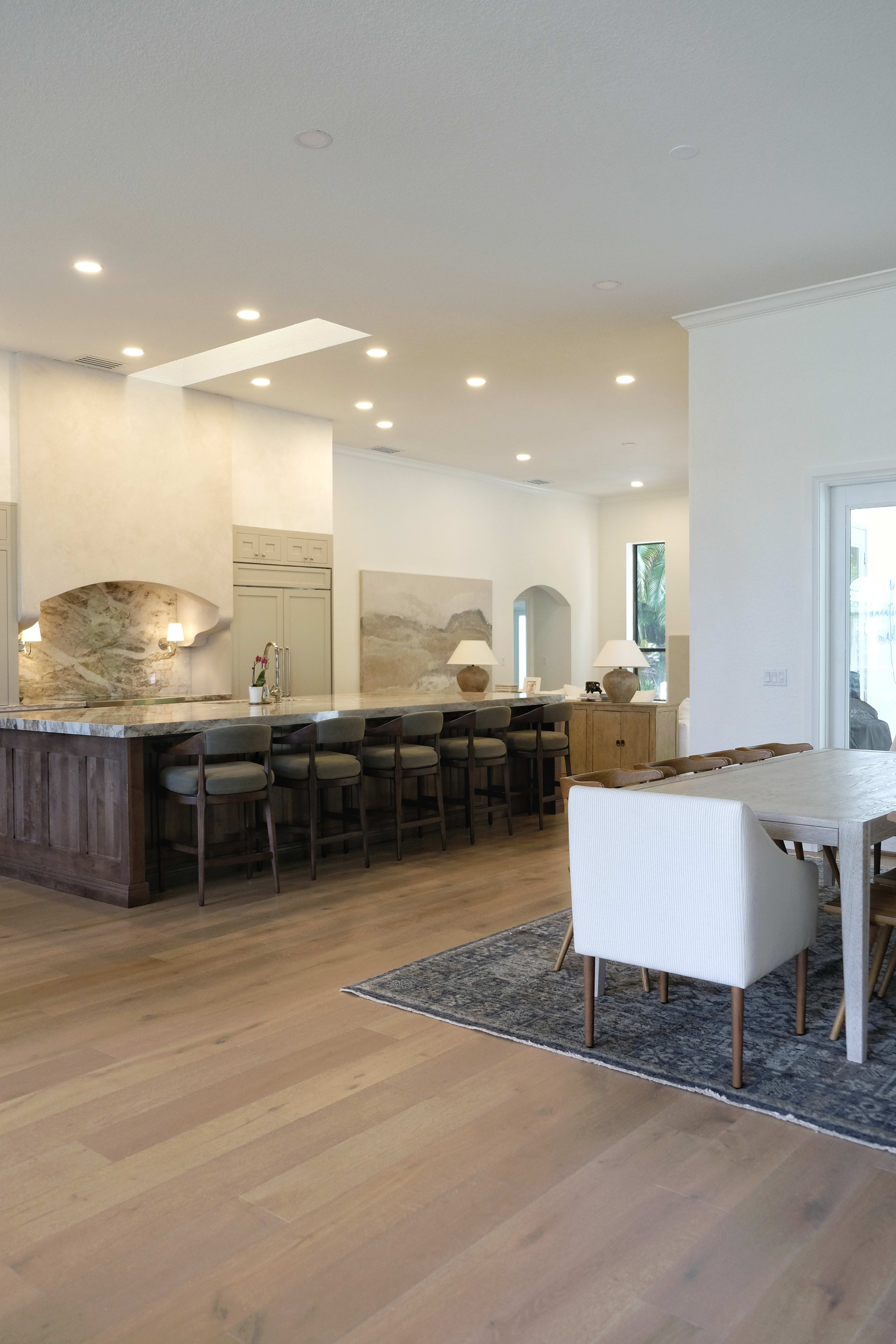 The image shows a modern kitchen and dining area with light wood flooring, a large island, and a dining table with upholstered chairs.