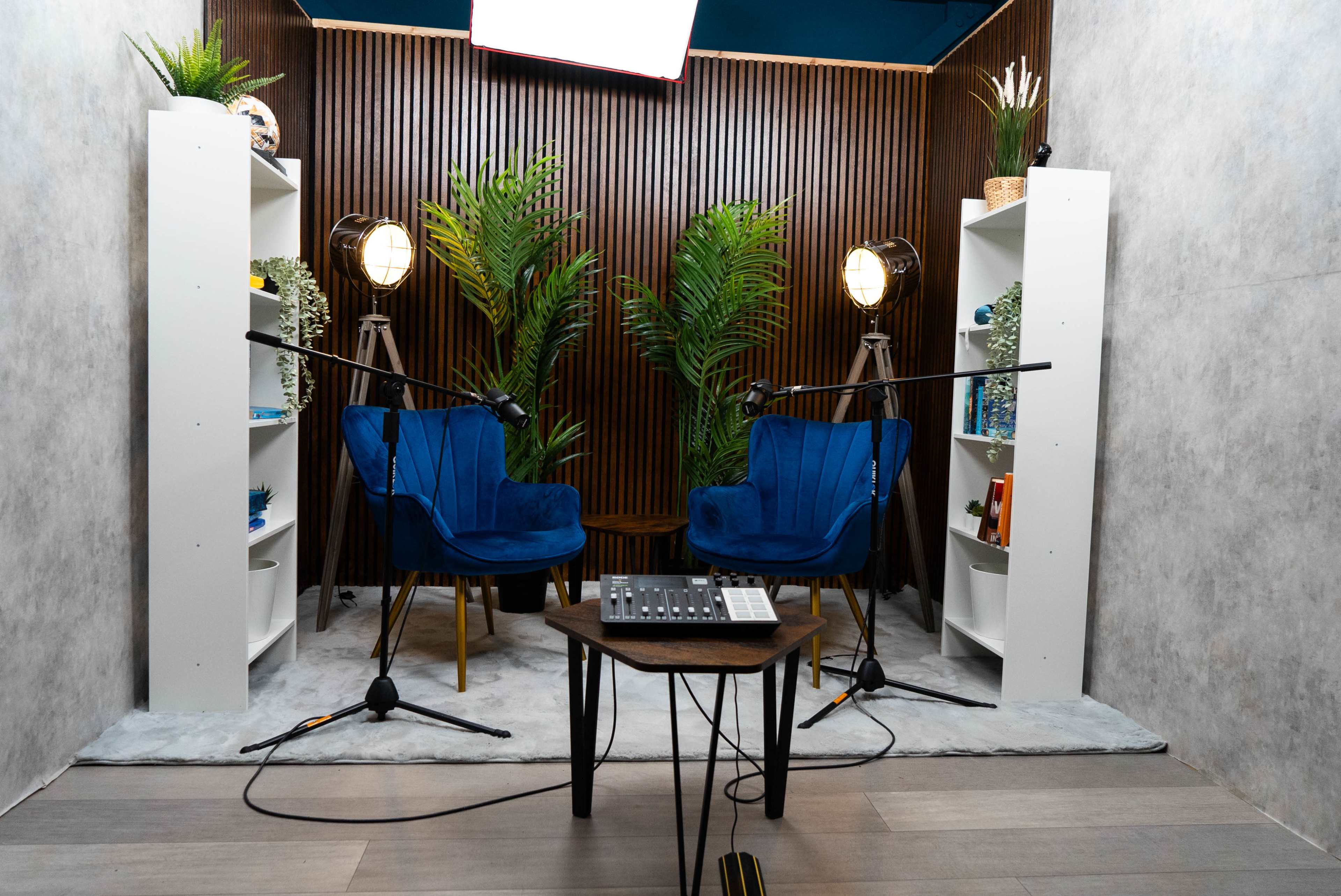 The scene features a podcast or interview setup with two blue chairs, microphones on stands, and a table with audio equipment, surrounded by greenery and decorative shelves.