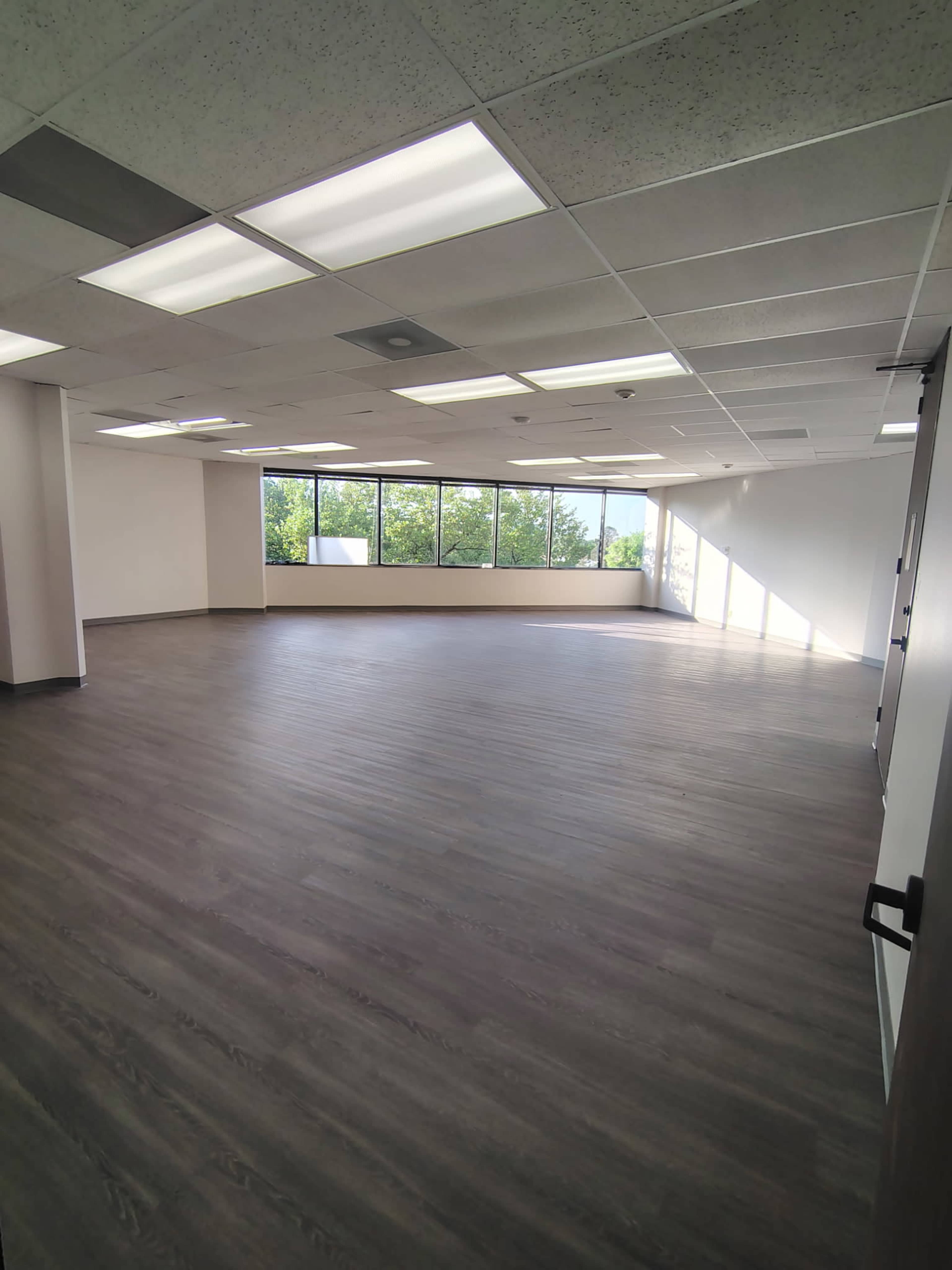 The image shows an empty office space with large windows and a smooth wooden floor.
