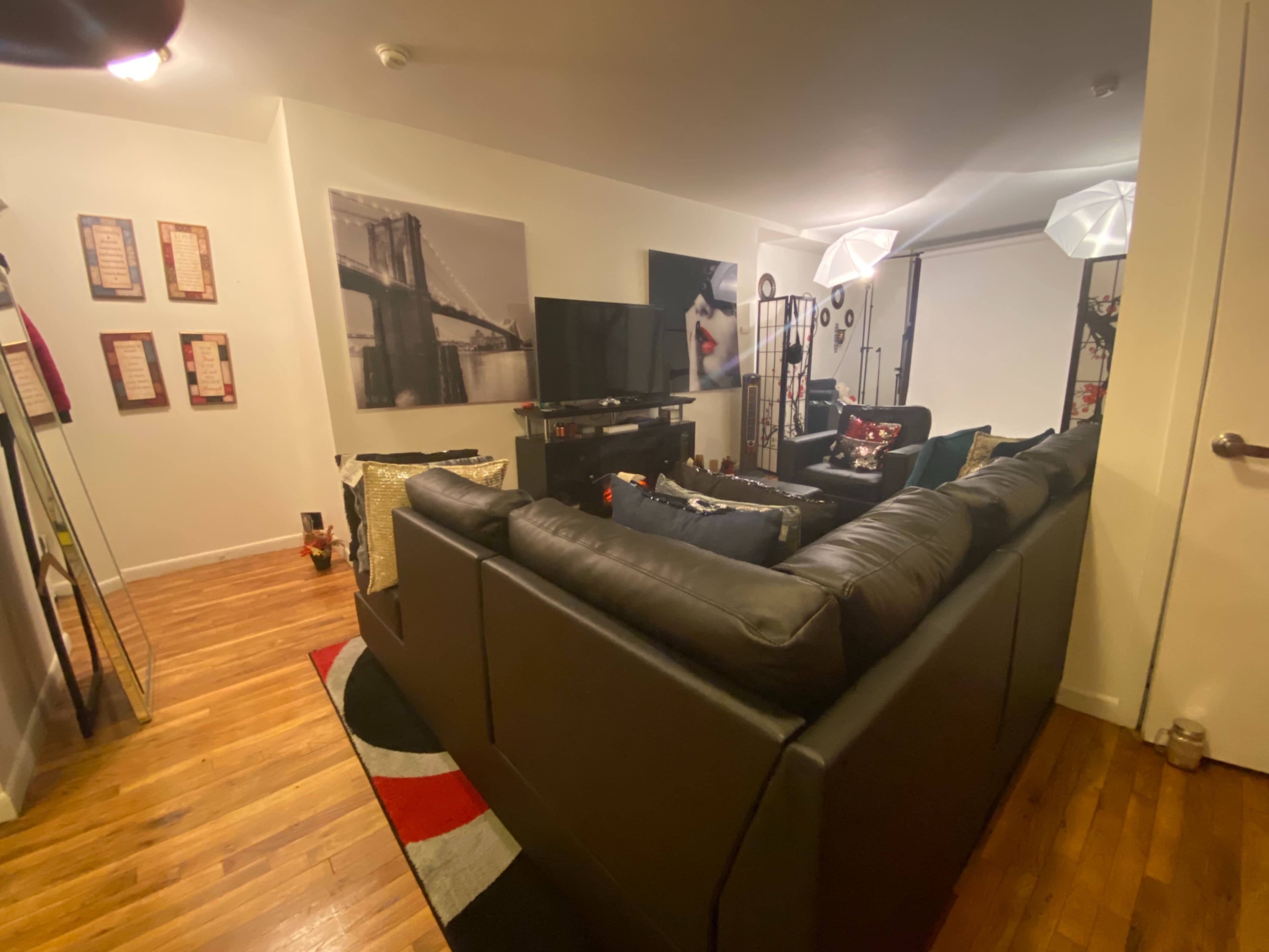 The image shows a living room with a sectional black sofa facing a television, framed art on the walls, and decorative lamps in the corner.