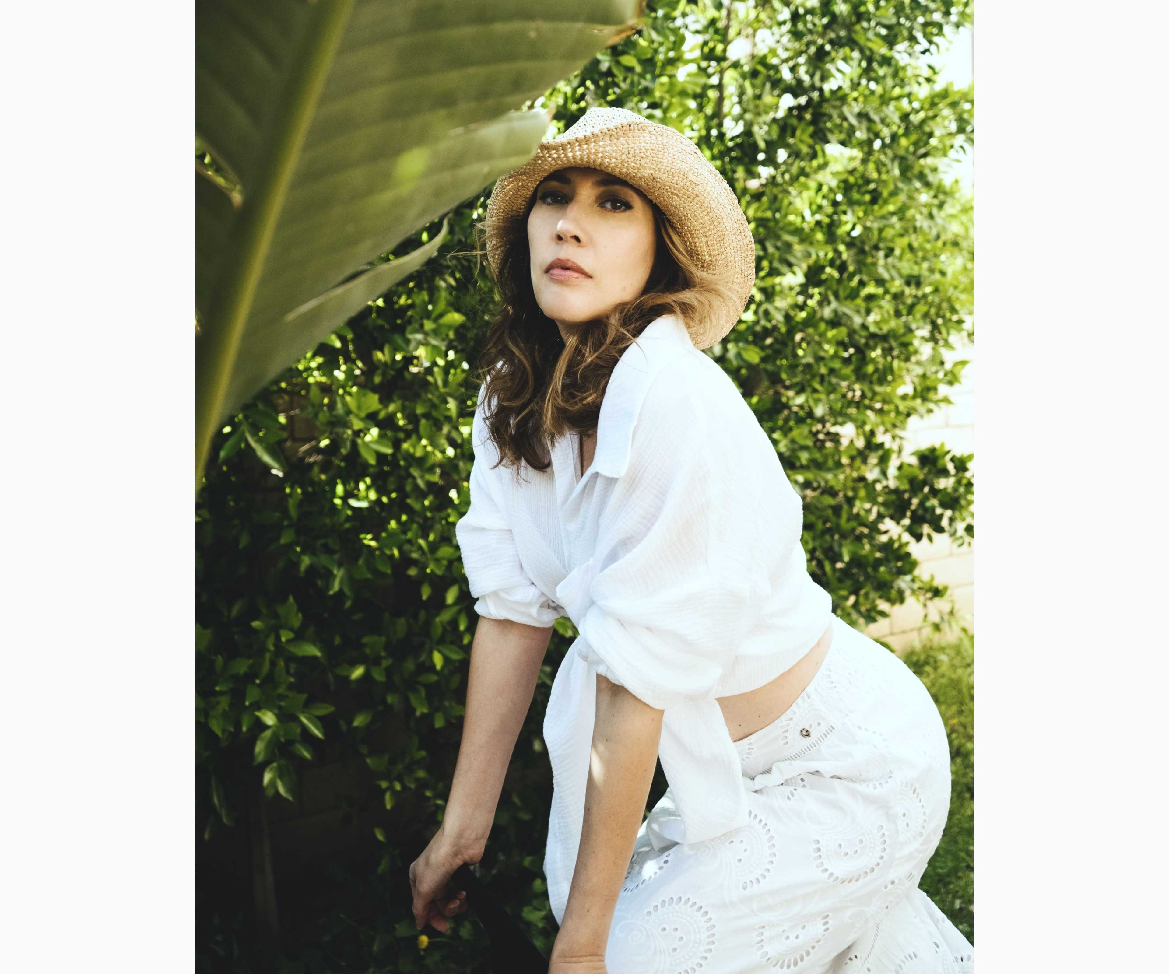 A woman in a white blouse and skirt poses among lush green foliage while wearing a straw hat.
