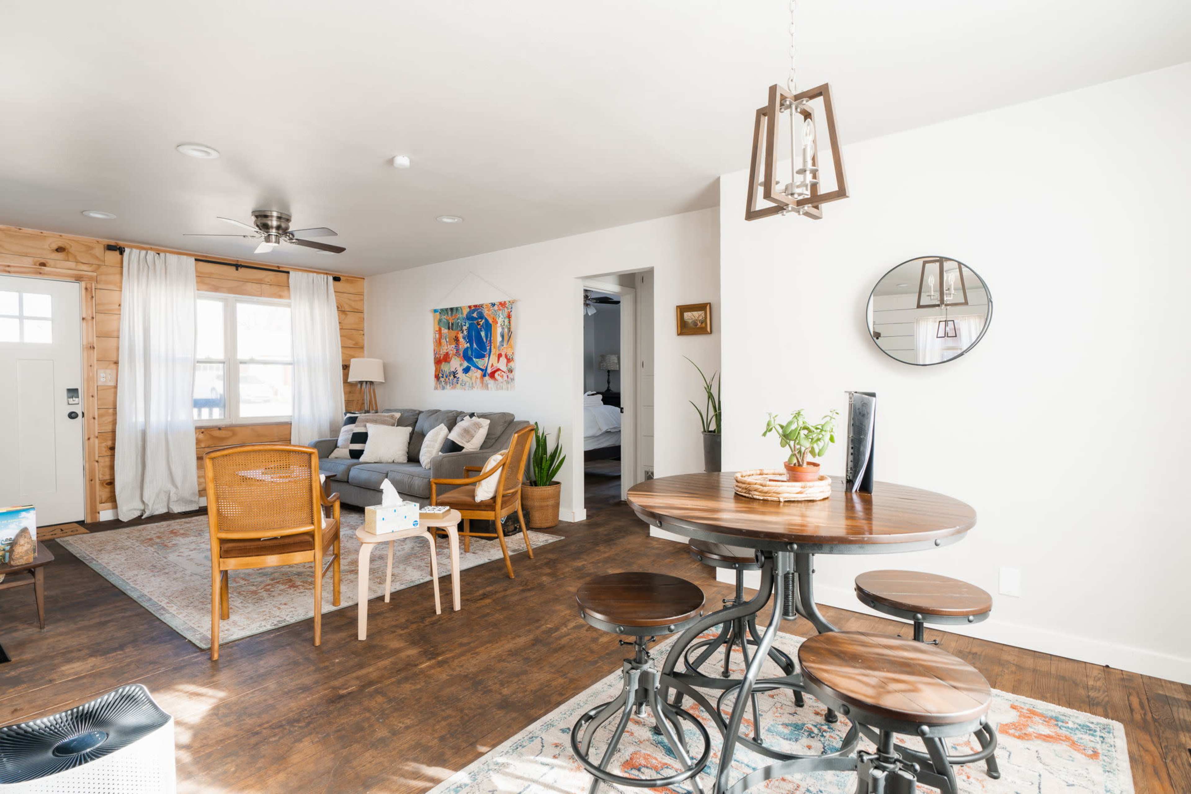 The image shows a cozy living space featuring a gray sofa, a wooden dining table with chairs, and a round mirror on the wall, along with a doorway leading to another room.