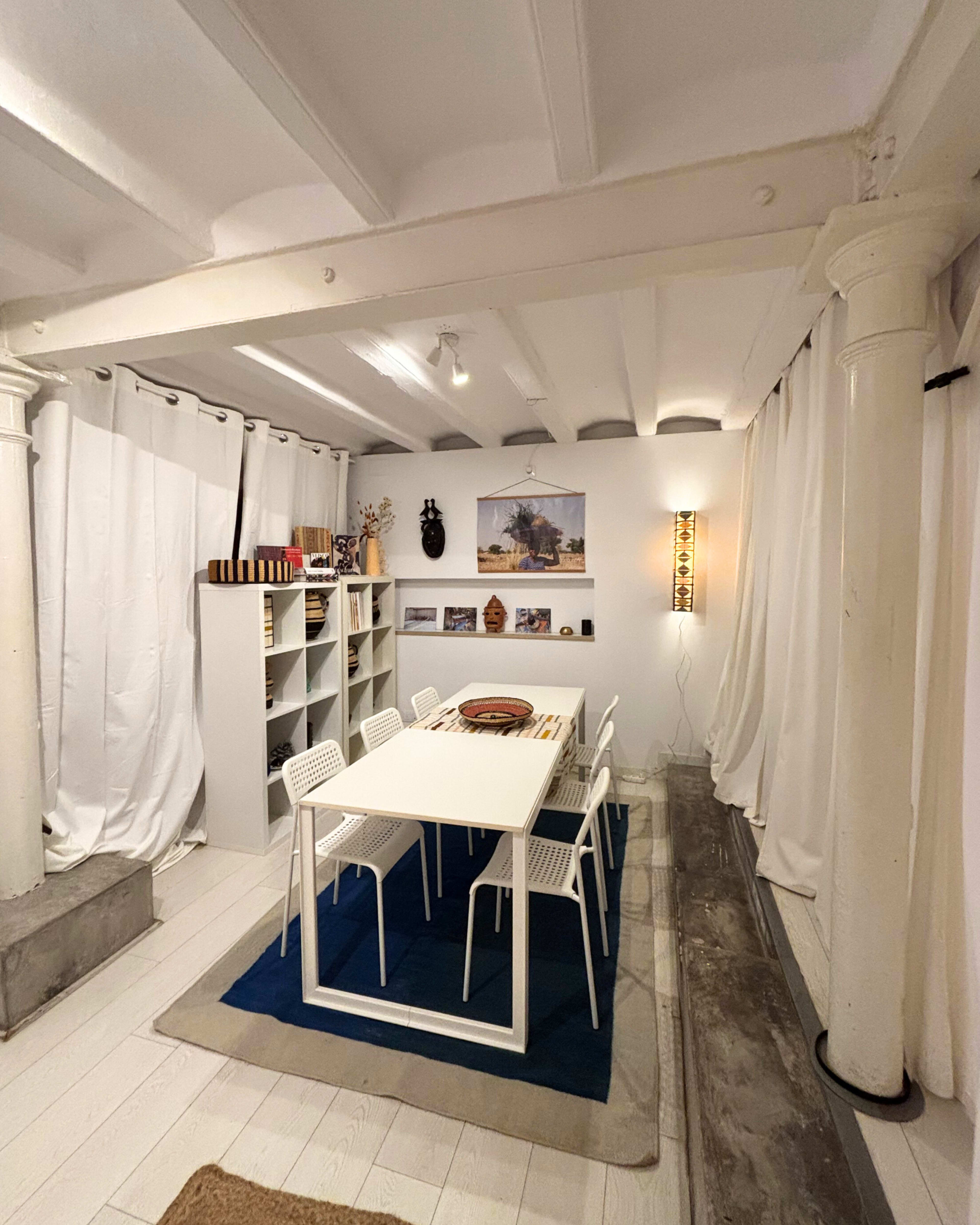 A spacious room features a white dining table surrounded by chairs, with sheer curtains, shelves, and decorative items lining the walls.