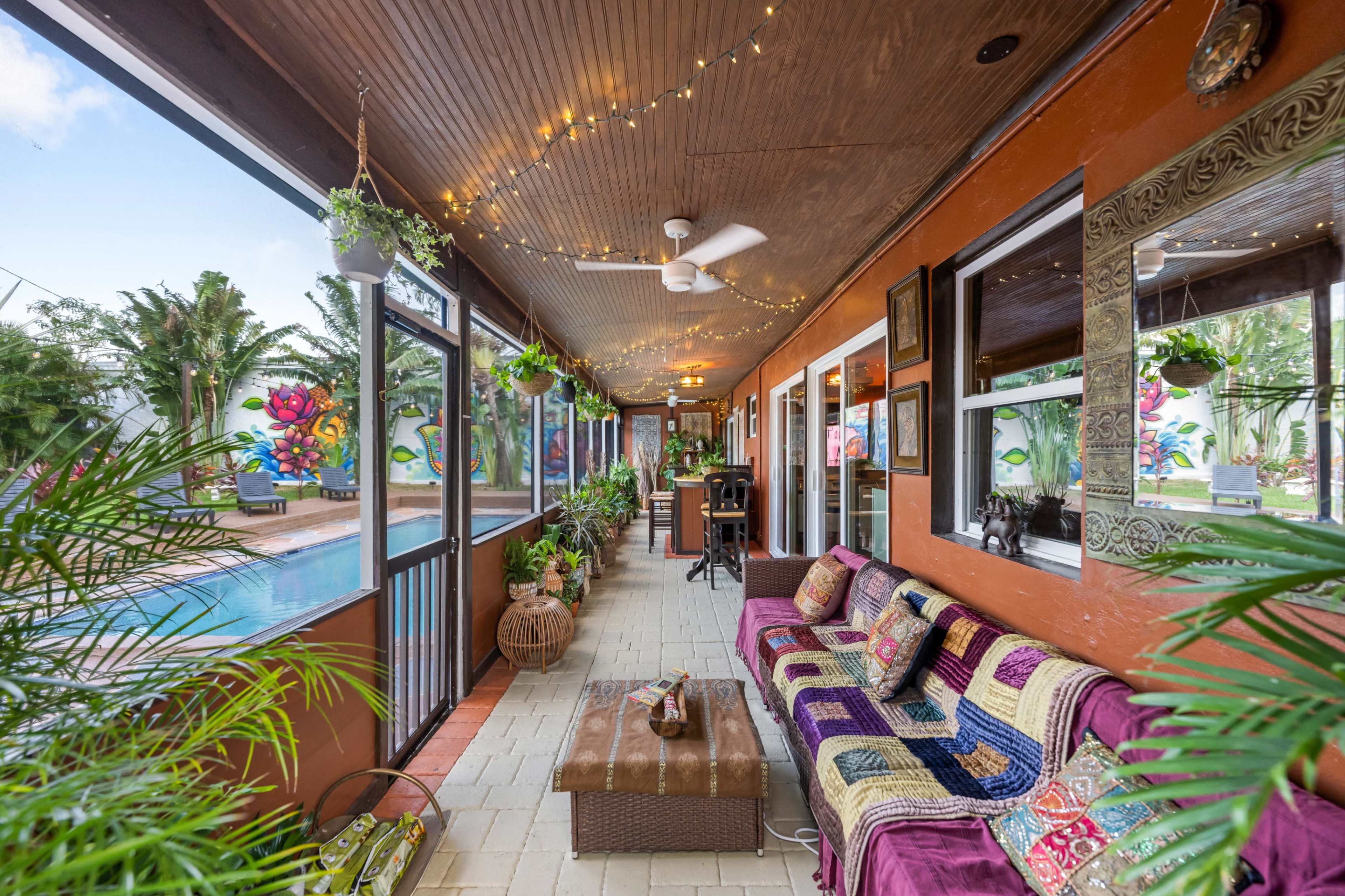 A covered porch with comfortable seating, lush plants, and a view of a swimming pool.