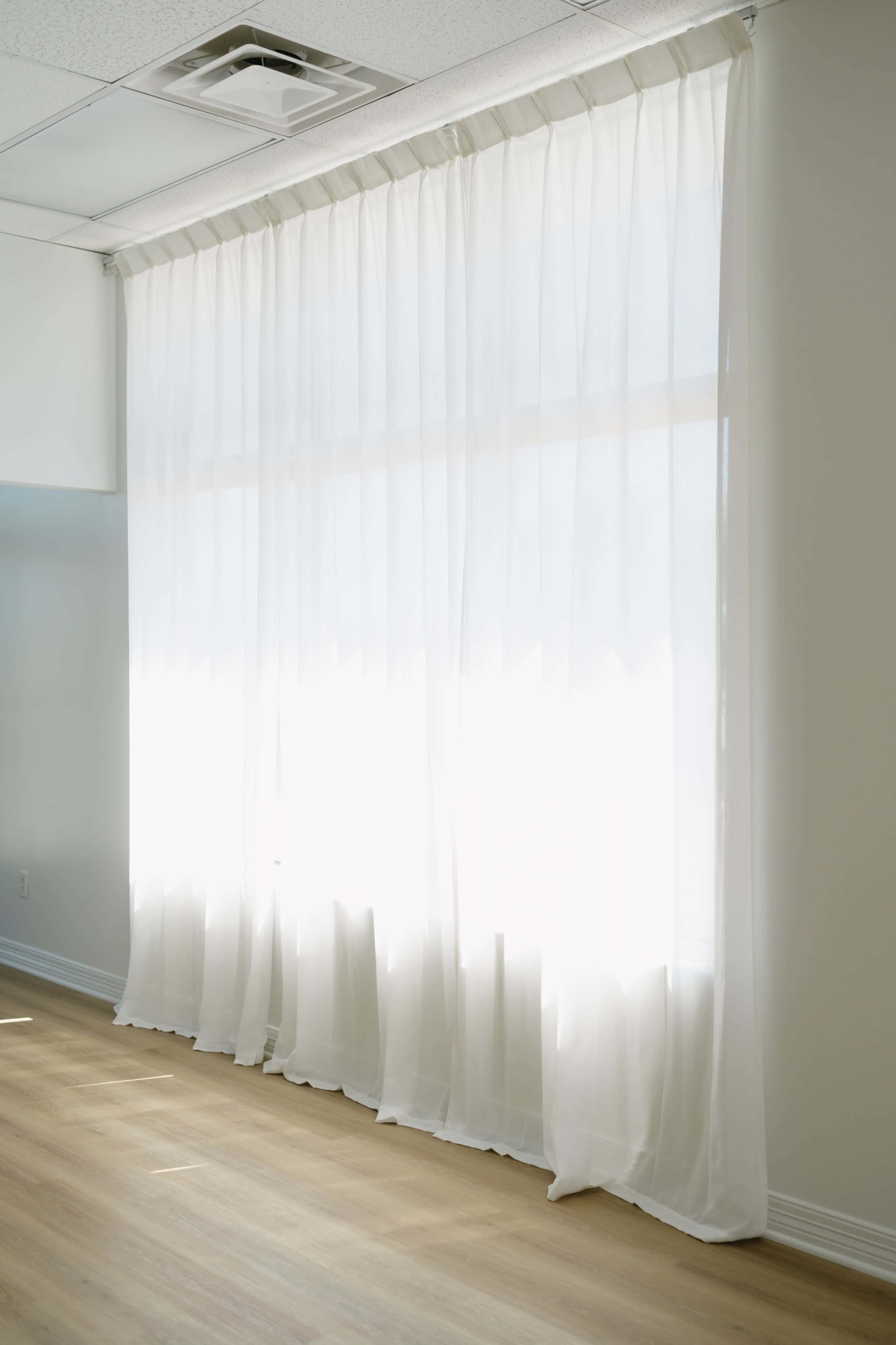 A sheer white curtain drapes over a large window, allowing natural light to filter into a room with light-colored wooden flooring.