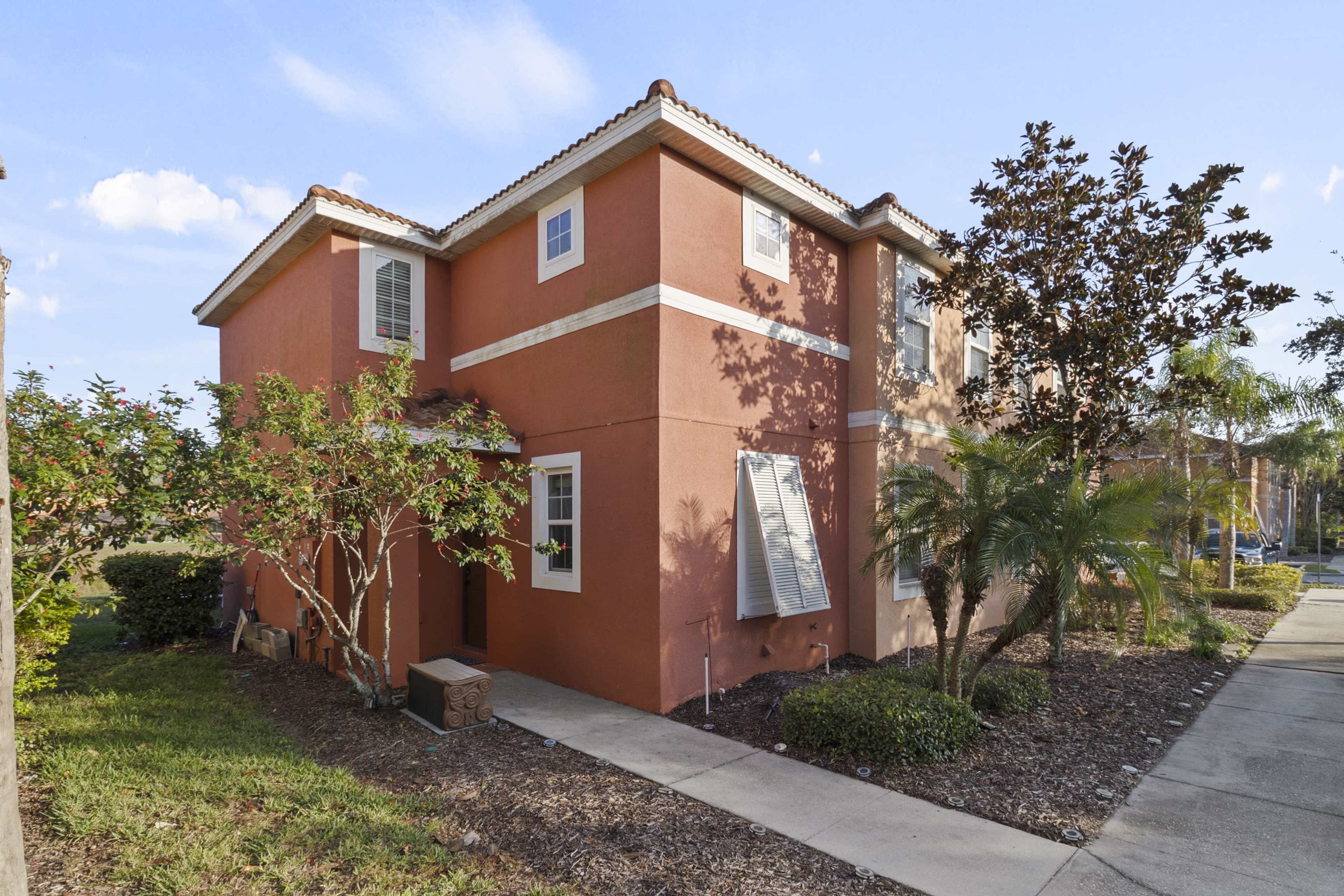 A two-story, coral-colored house with white trim is surrounded by landscaped shrubs and trees.