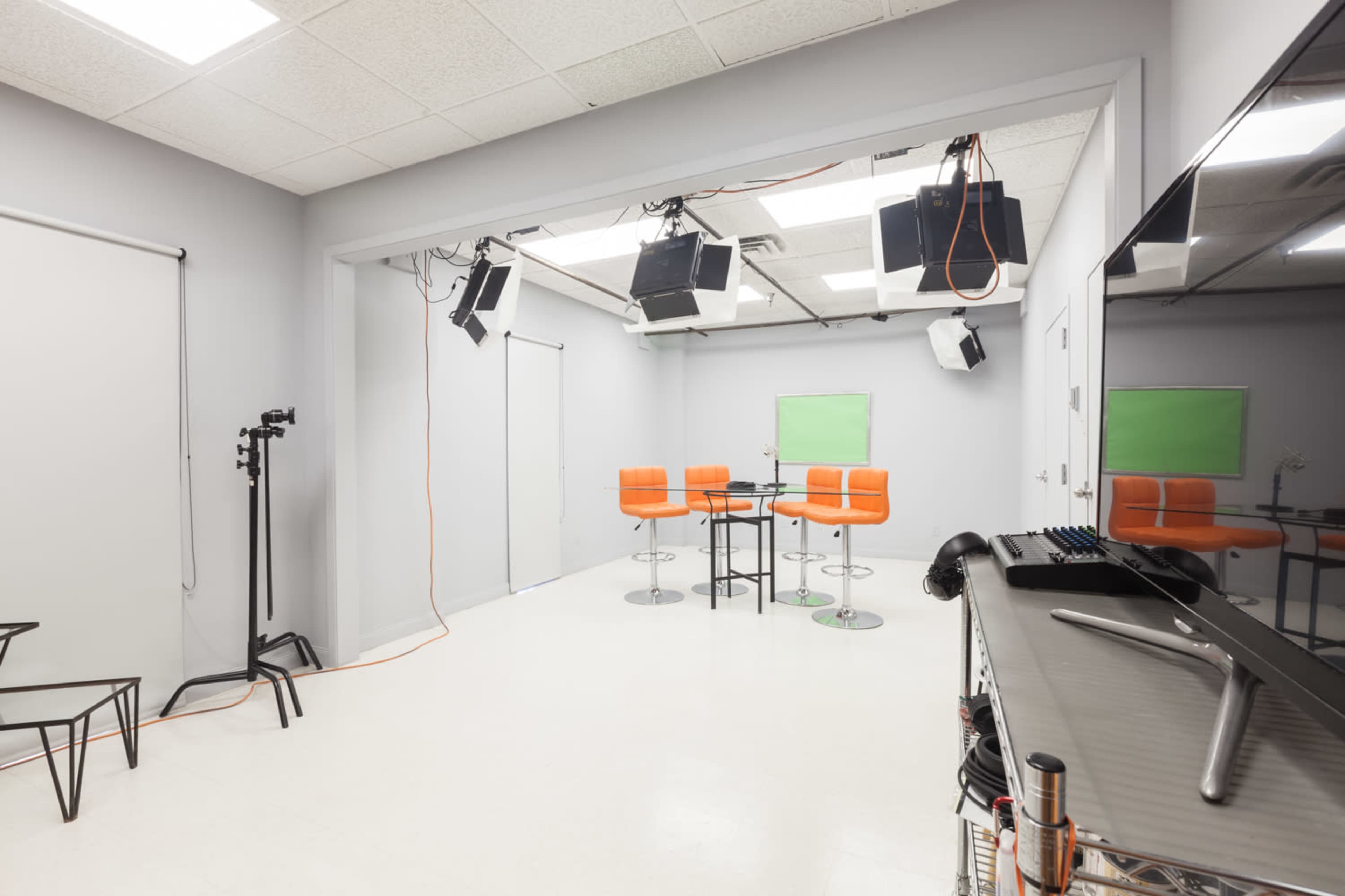A modern studio space features orange bar stools around a black table, with professional lighting equipment and green screen areas.