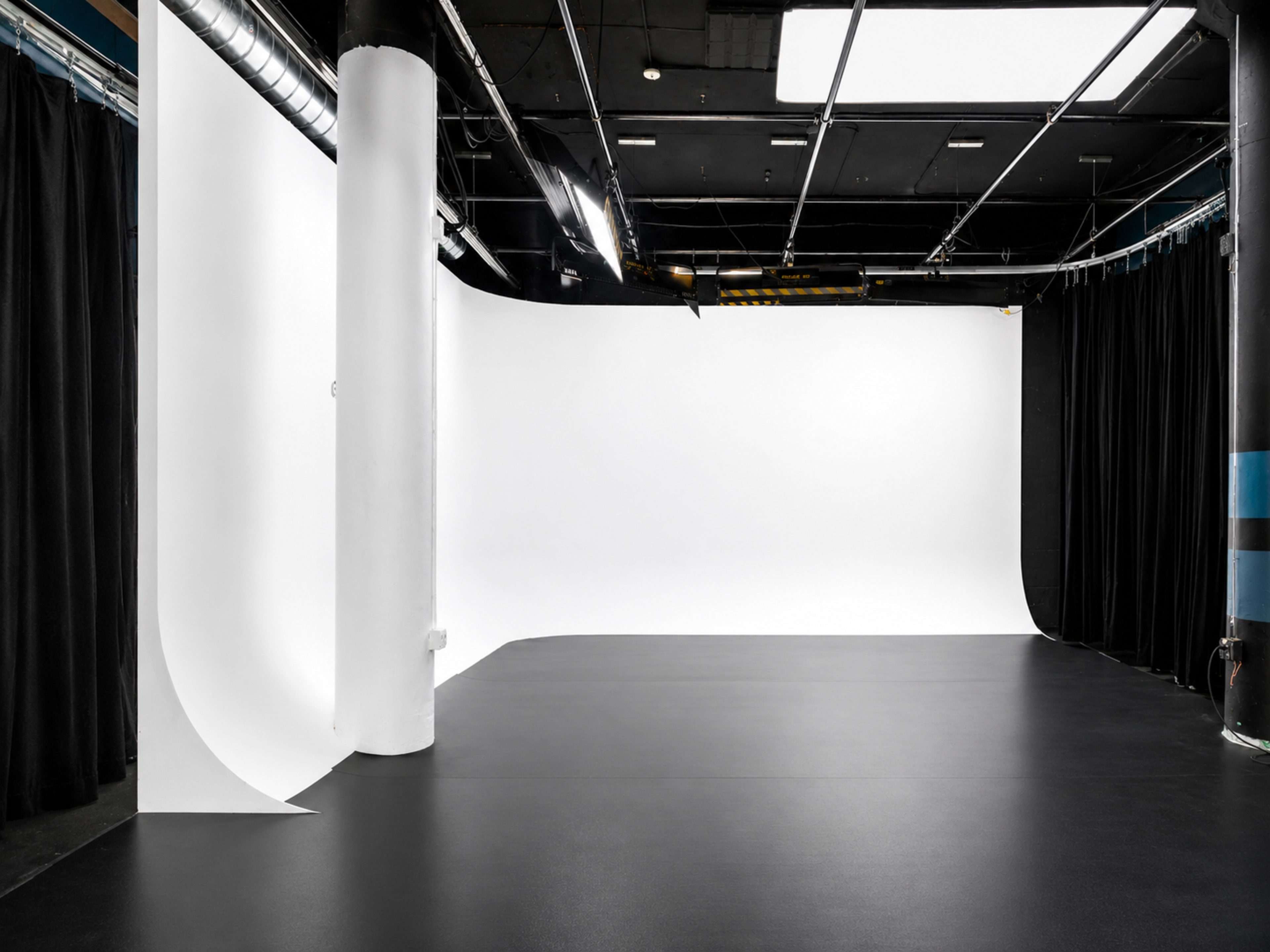 The image shows a spacious studio with a black floor, white backdrop, and black curtains draped along the sides.