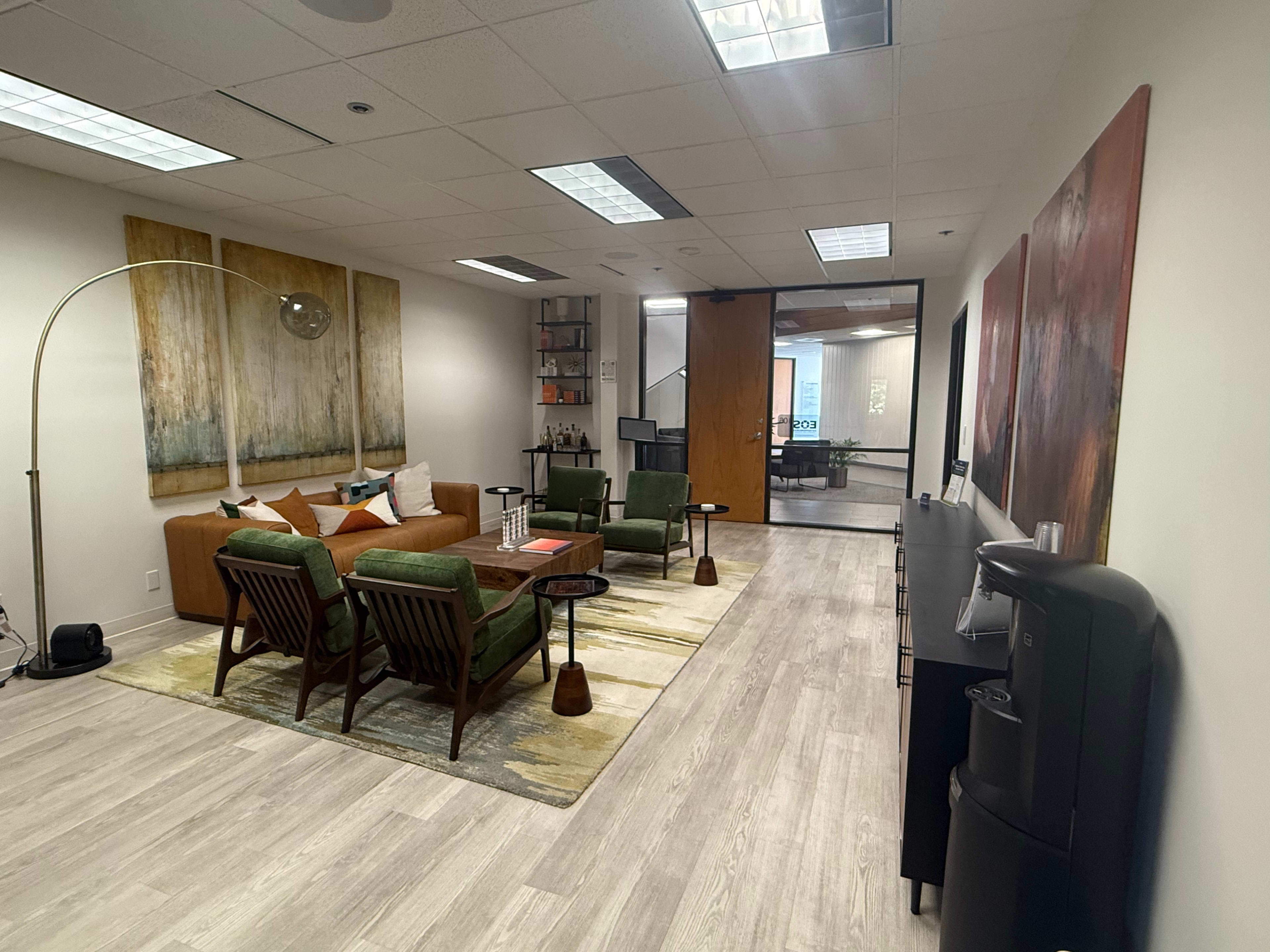 The image shows a modern office lounge area with a brown sofa, green chairs, a large area rug, and a coffee station along one wall.
