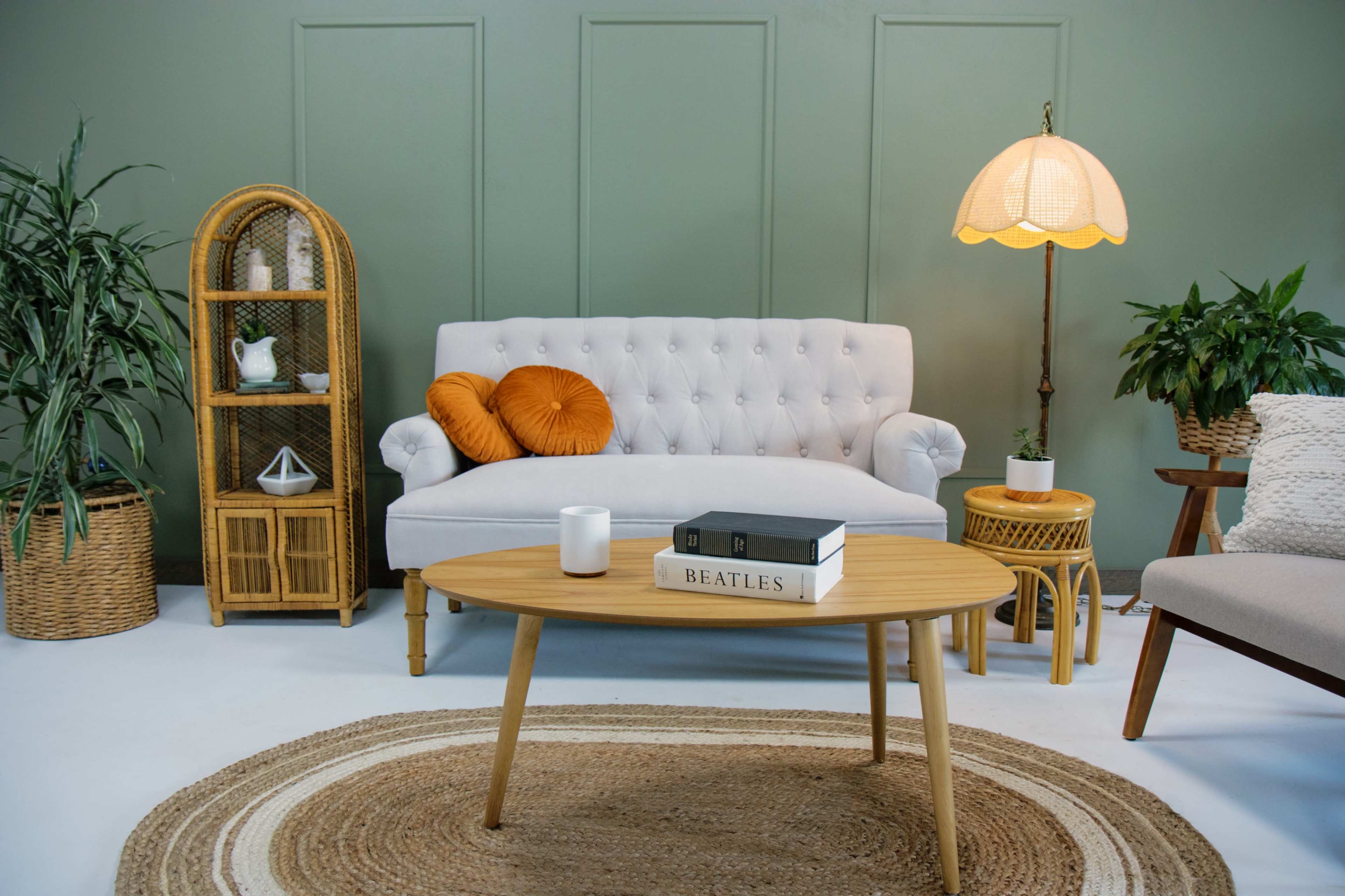 A light grey tufted sofa sits in a green room alongside a round woven rug, a wooden coffee table with books, and decorative plants.
