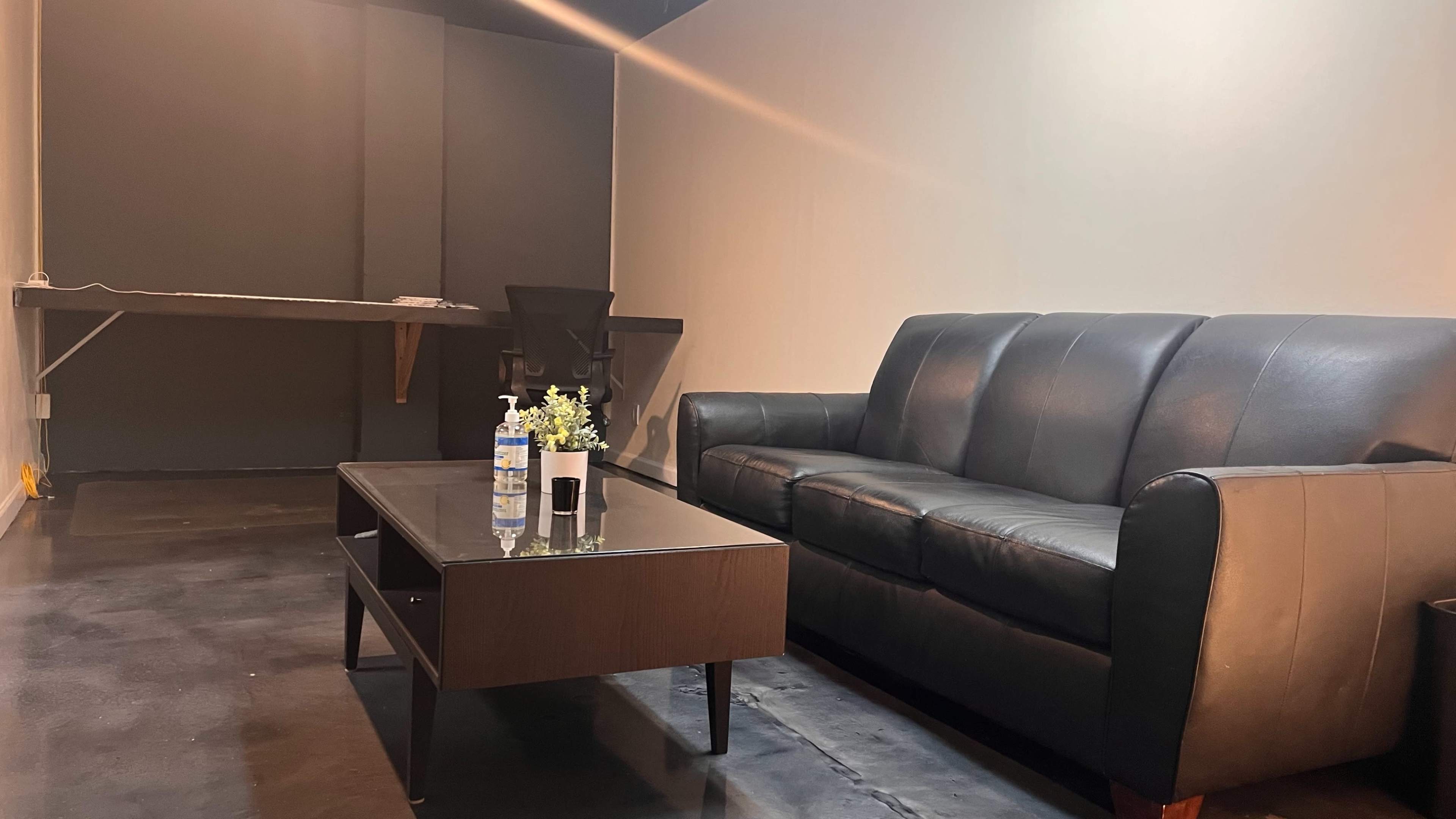 A minimalistic interior with a black leather couch, a coffee table, and a desk with a chair against a gray wall.