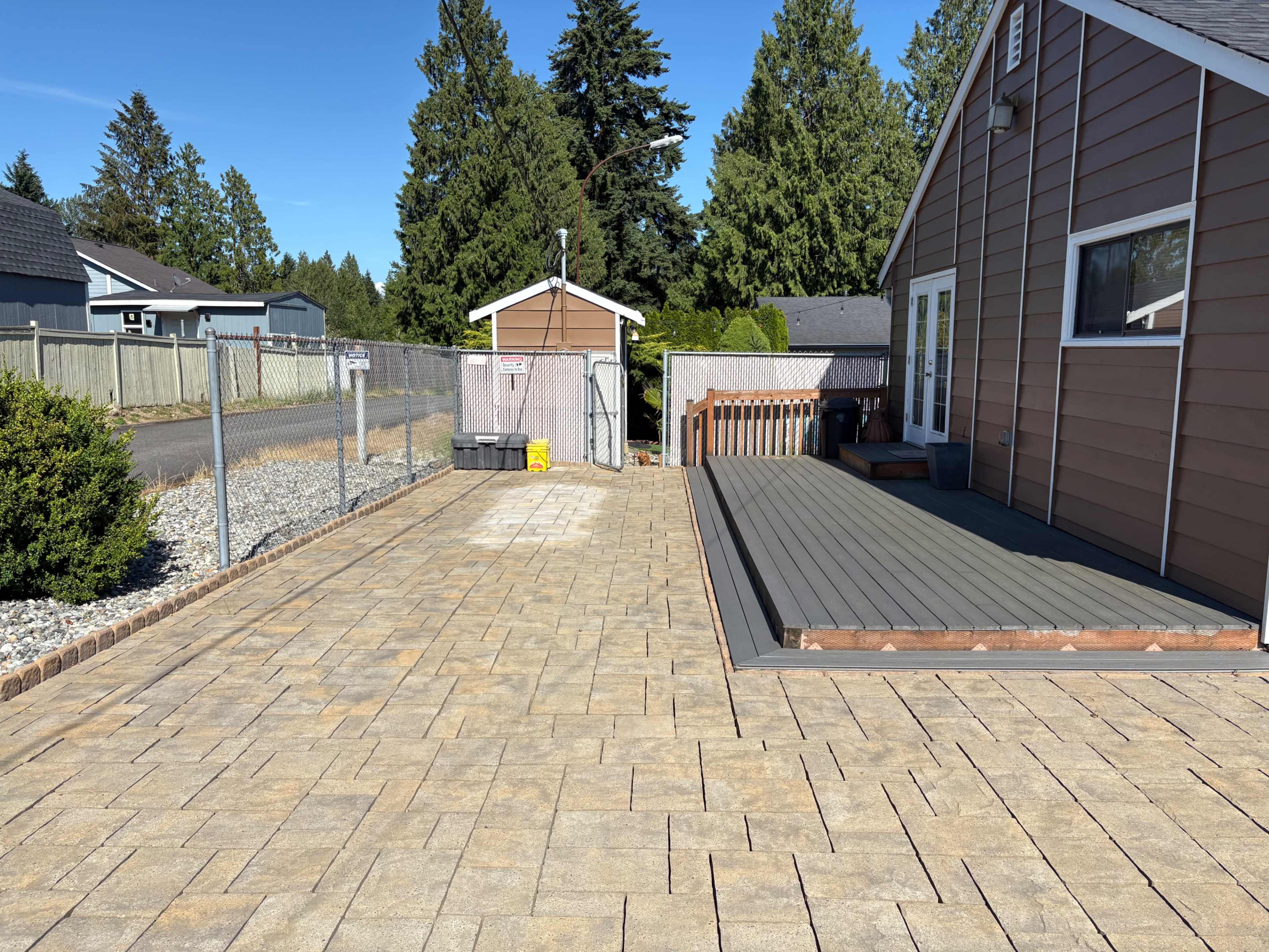 The image shows a landscaped area featuring a paved patio or driveway, bordered by a wooden deck and surrounded by trees and a fence.