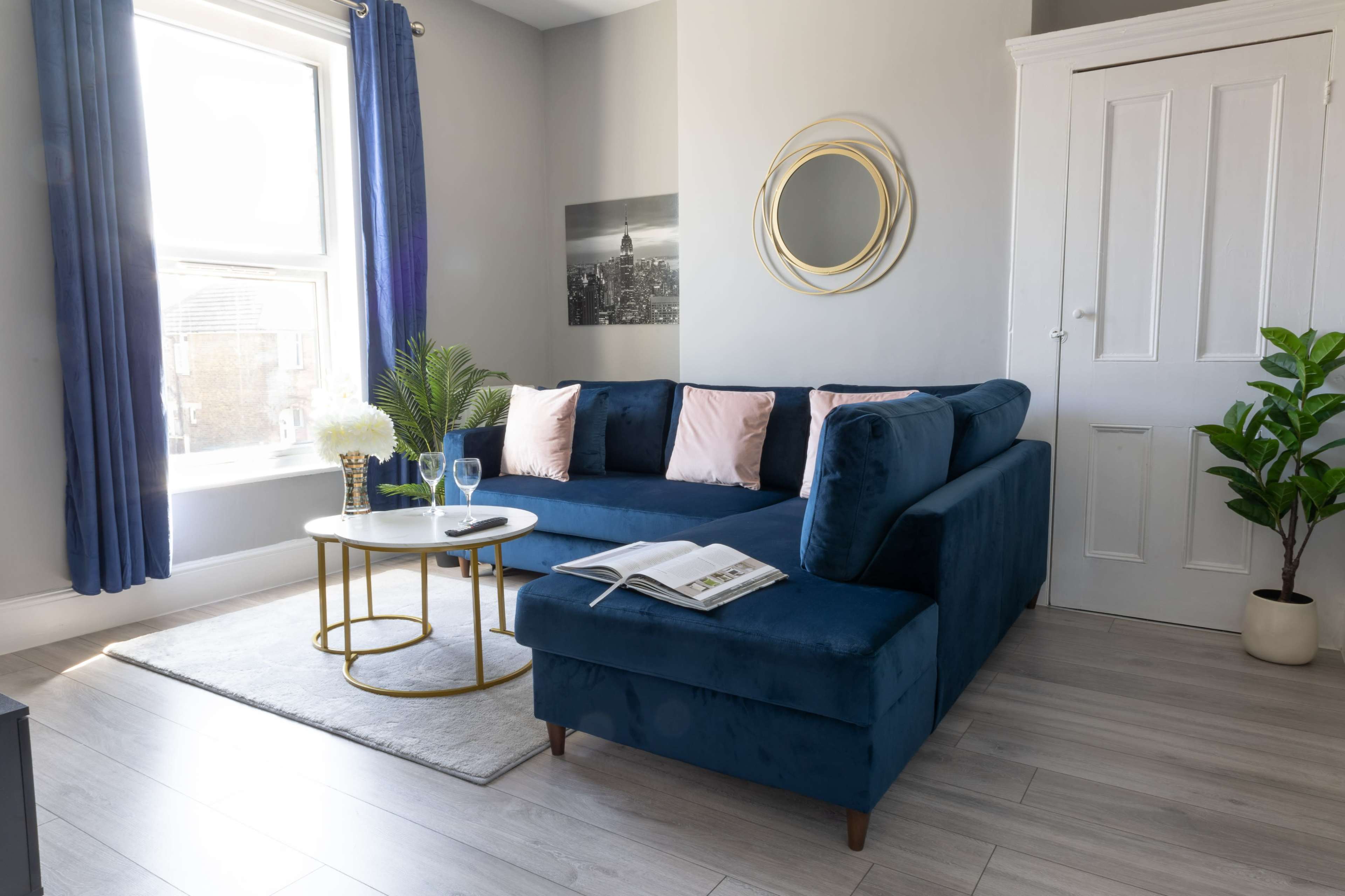 A blue sectional sofa with pink pillows is positioned next to a coffee table in a well-lit living room featuring a large window and a houseplant.
