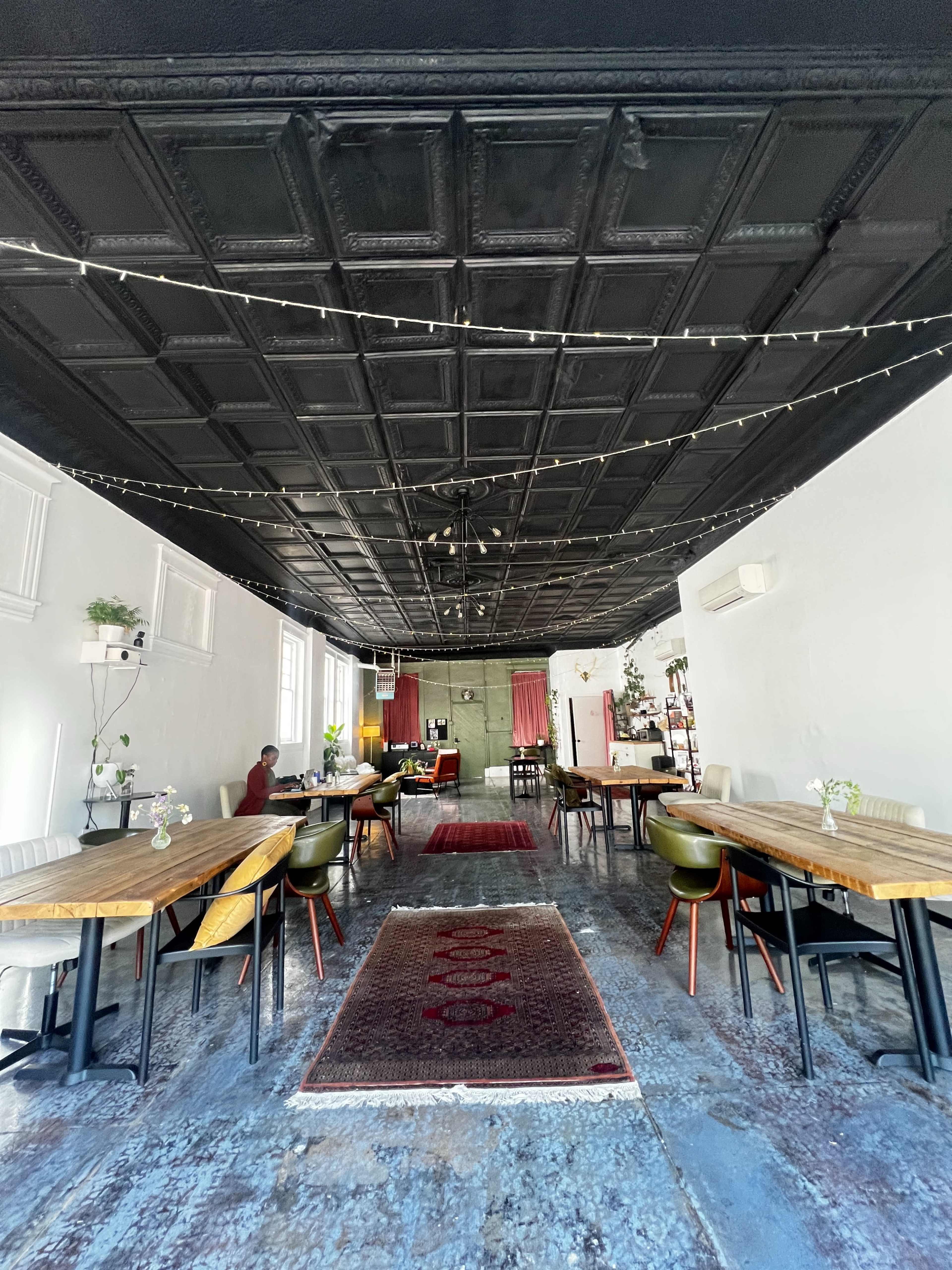 The image shows a spacious dining area with wooden tables and chairs, a patterned rug on the floor, and a dark ceiling with string lights.