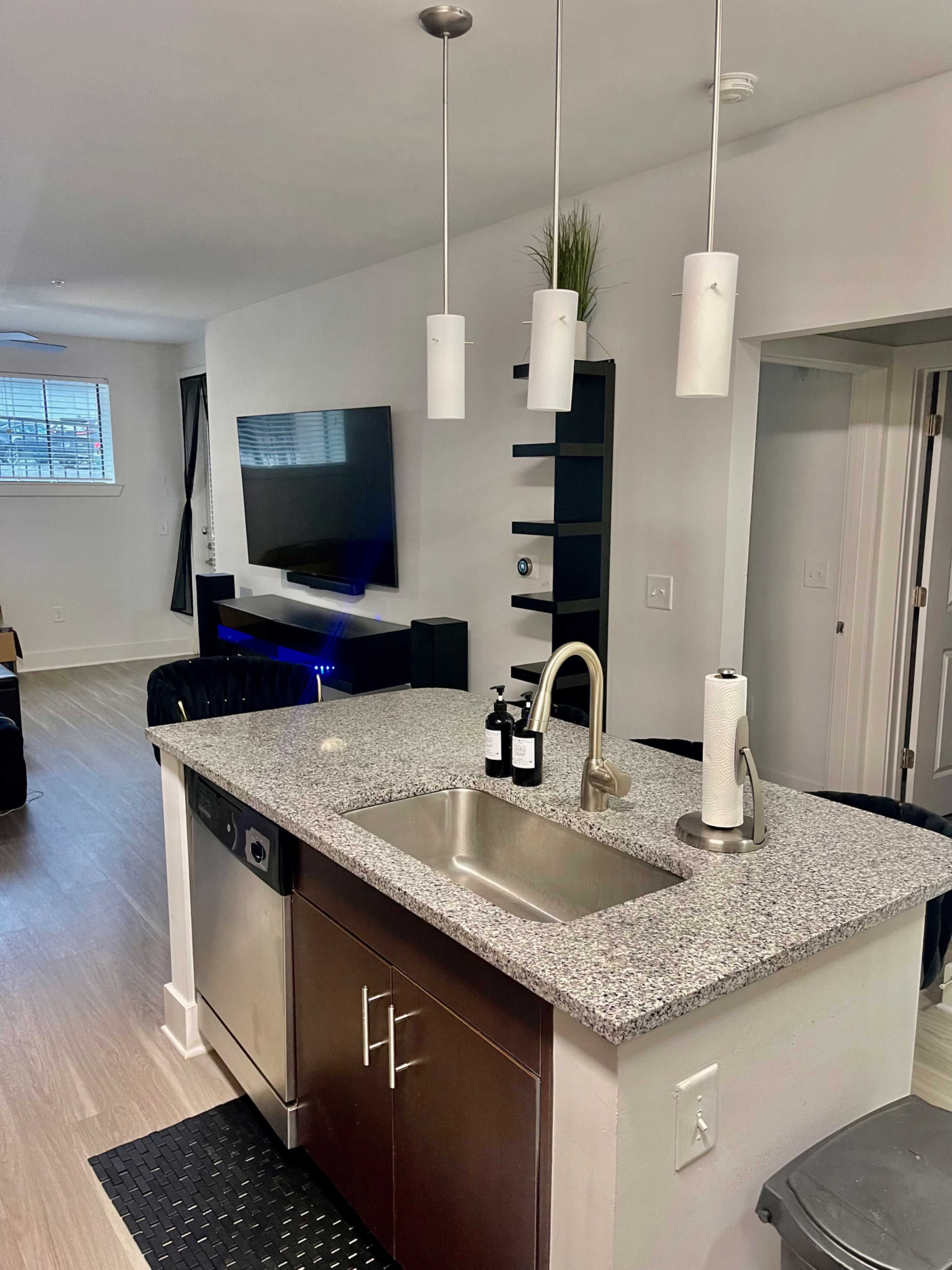 A modern kitchen features a granite countertop with a sink, a dishwasher, and pendant lights, while a living area is visible in the background with a TV and seating.