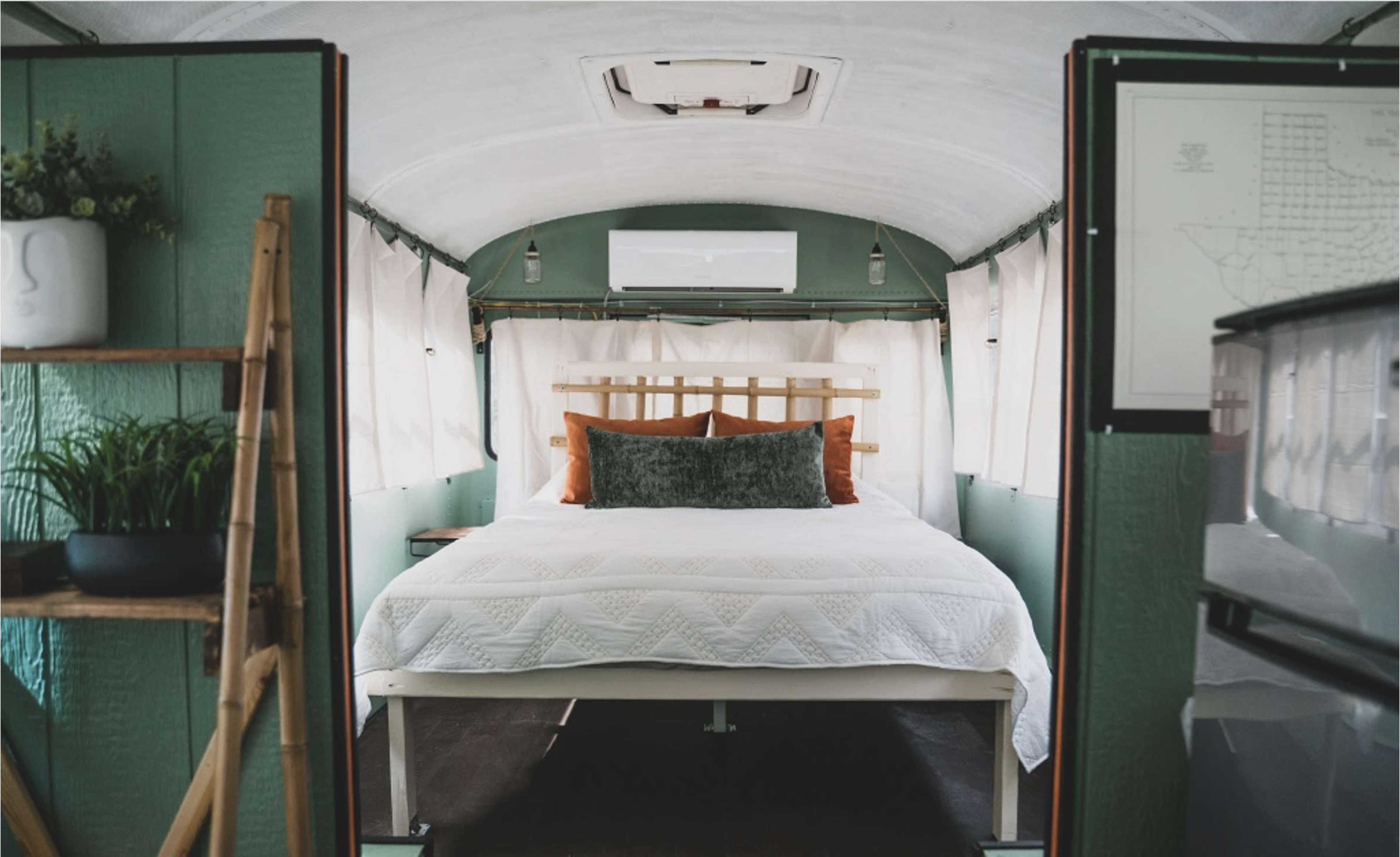 The interior of a converted bus featuring a bed with decorative pillows, surrounded by green walls and large windows.