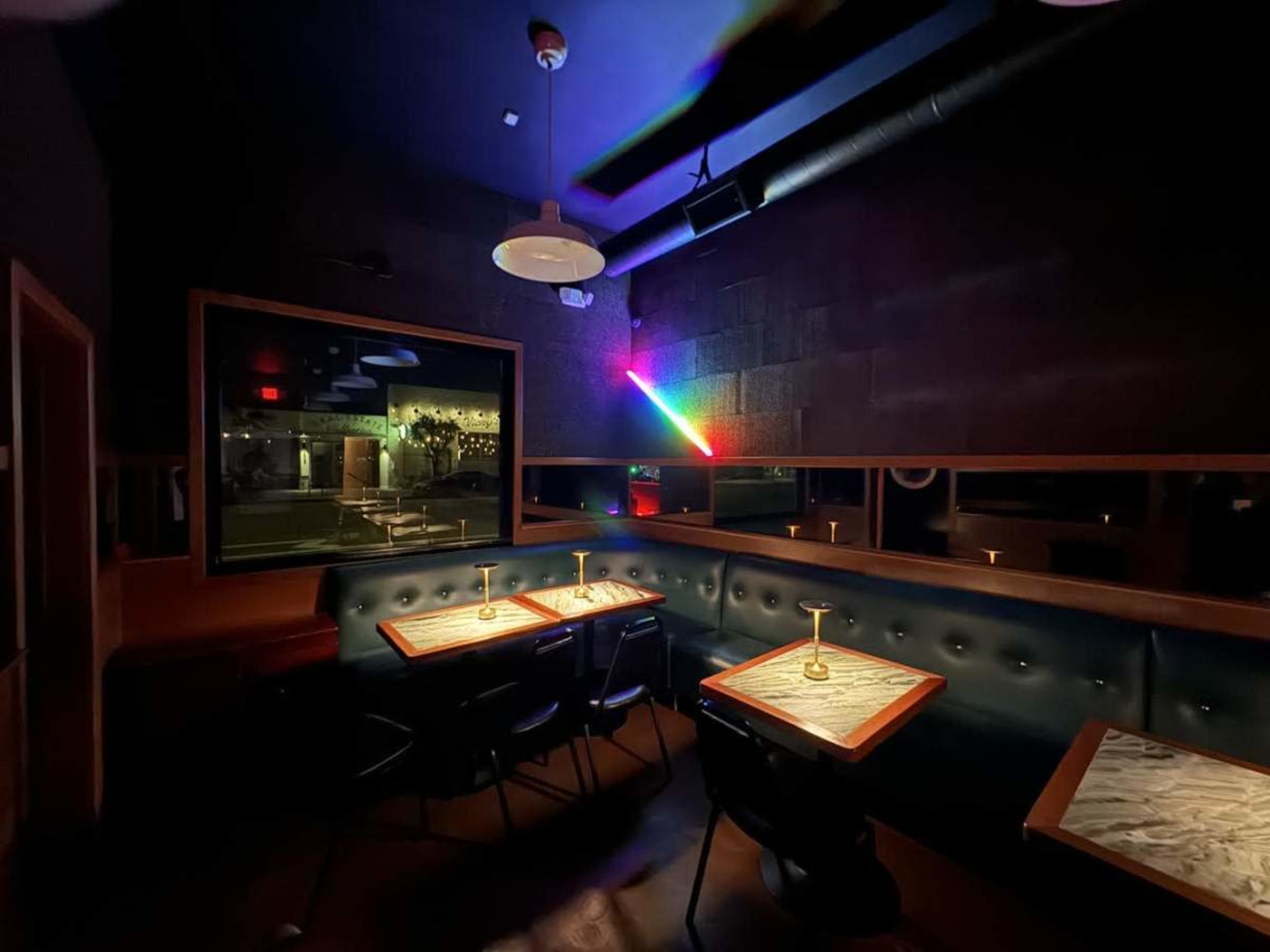 The image shows a dimly lit bar with green-velvet benches, wooden tables, and a colorful rainbow light projection on the wall.