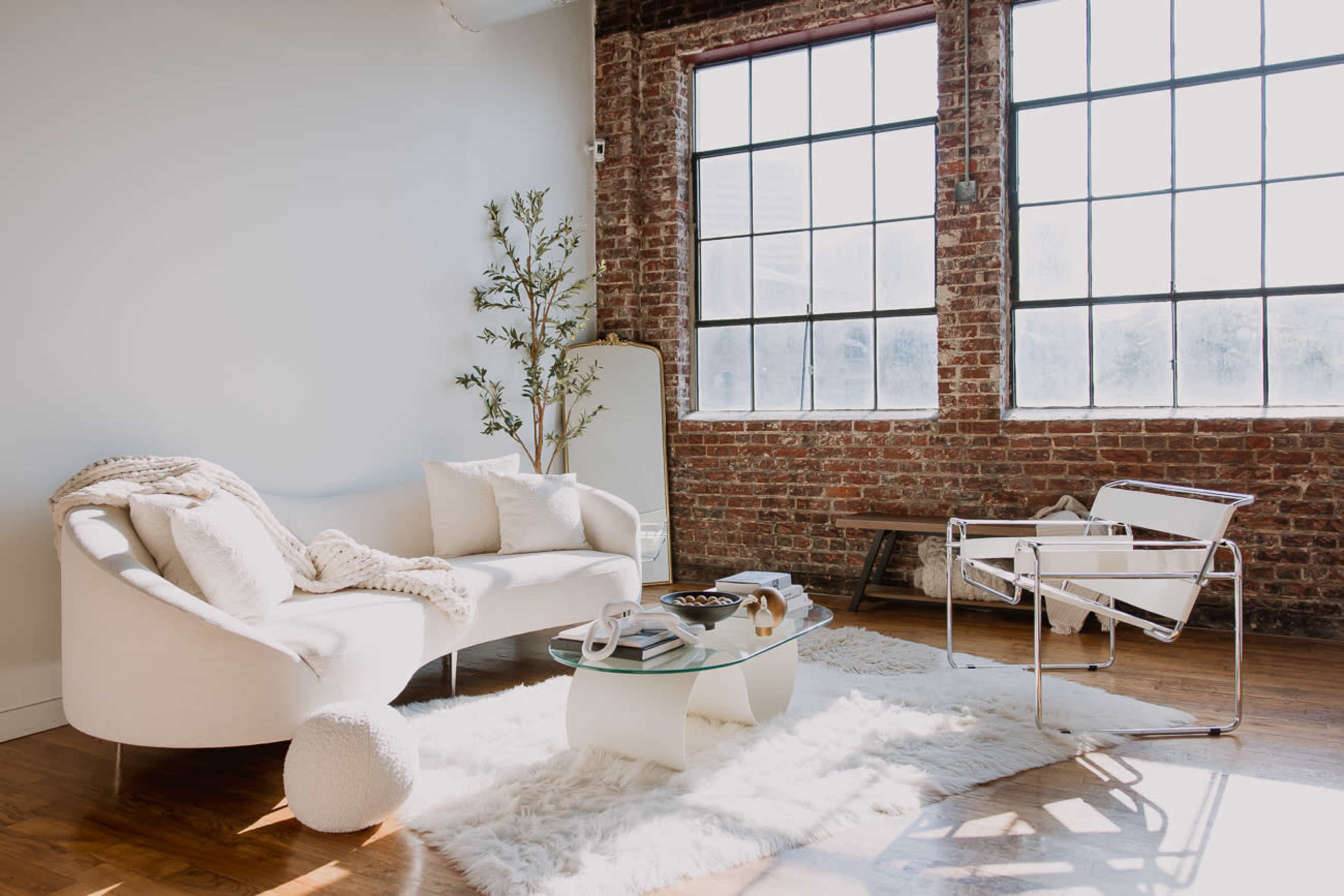 A bright living space features a white sofa and chair set, a glass coffee table, and large windows framed by exposed brick walls.