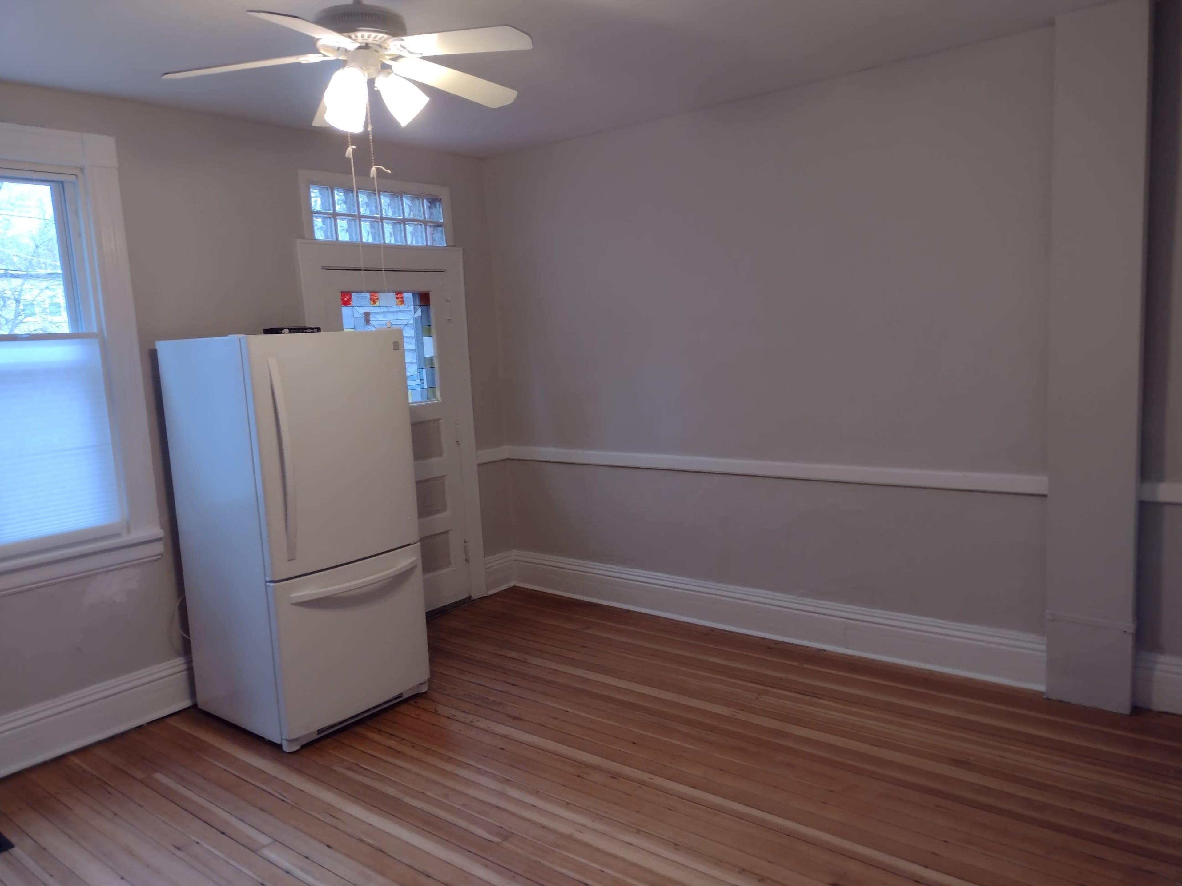 A spacious room with wooden floors, a refrigerator in the corner, and a ceiling fan.