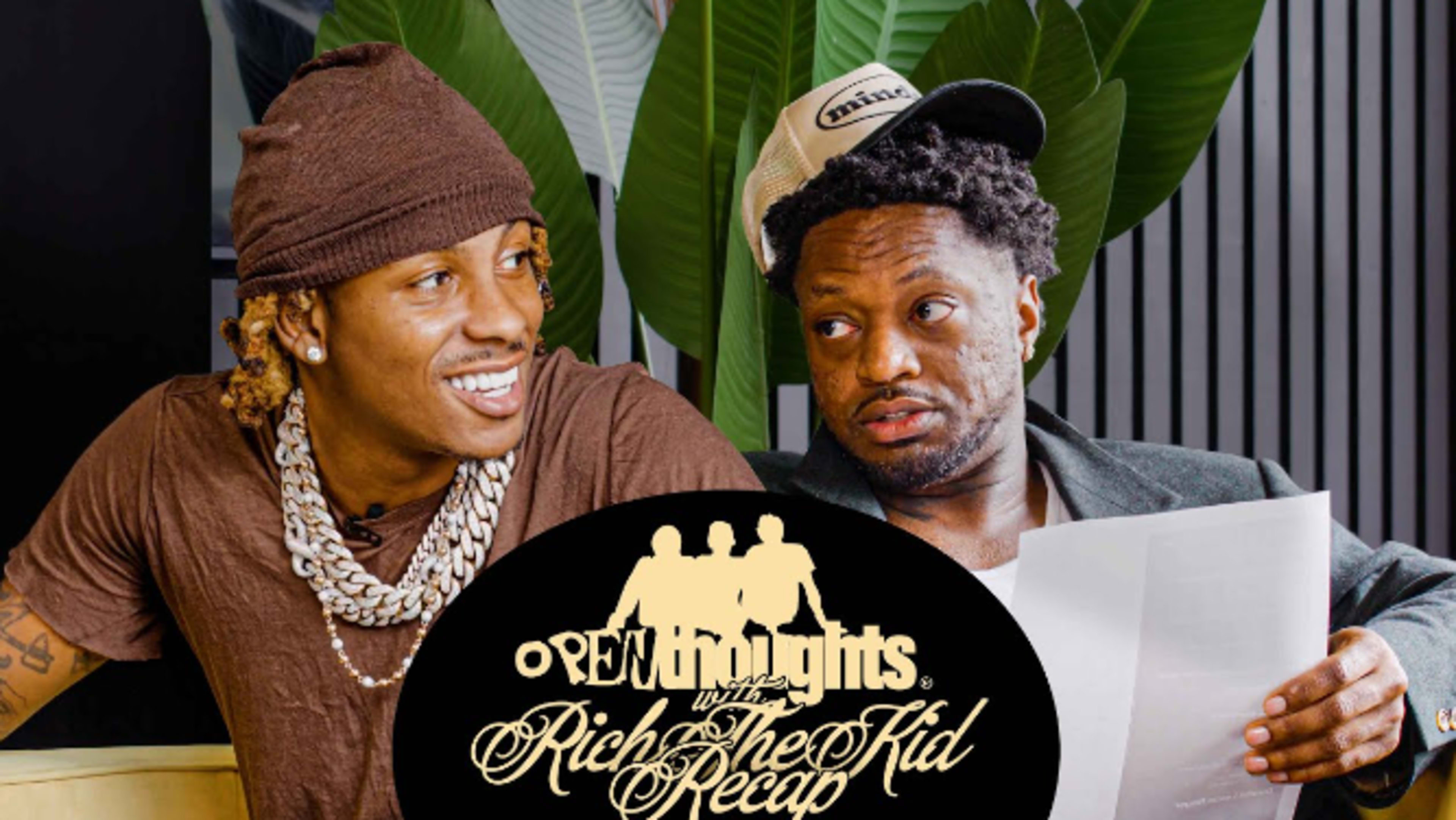 Two men sit together, one wearing a brown beanie and large necklace, while the other, in a jacket and cap, holds papers, both surrounded by indoor plants.