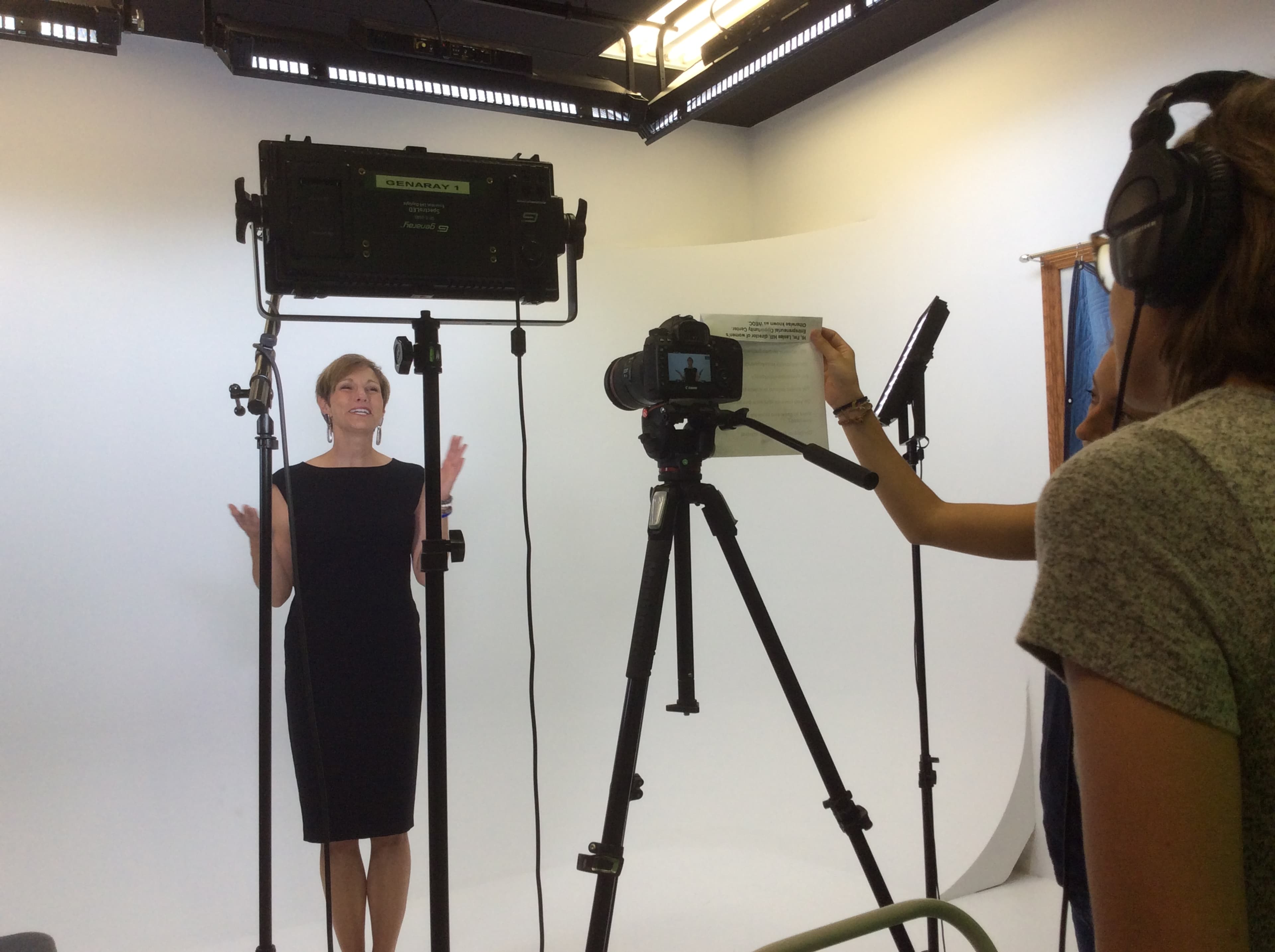 A woman stands in front of a camera and lights while holding a script, as another person operates the camera in a studio setting.