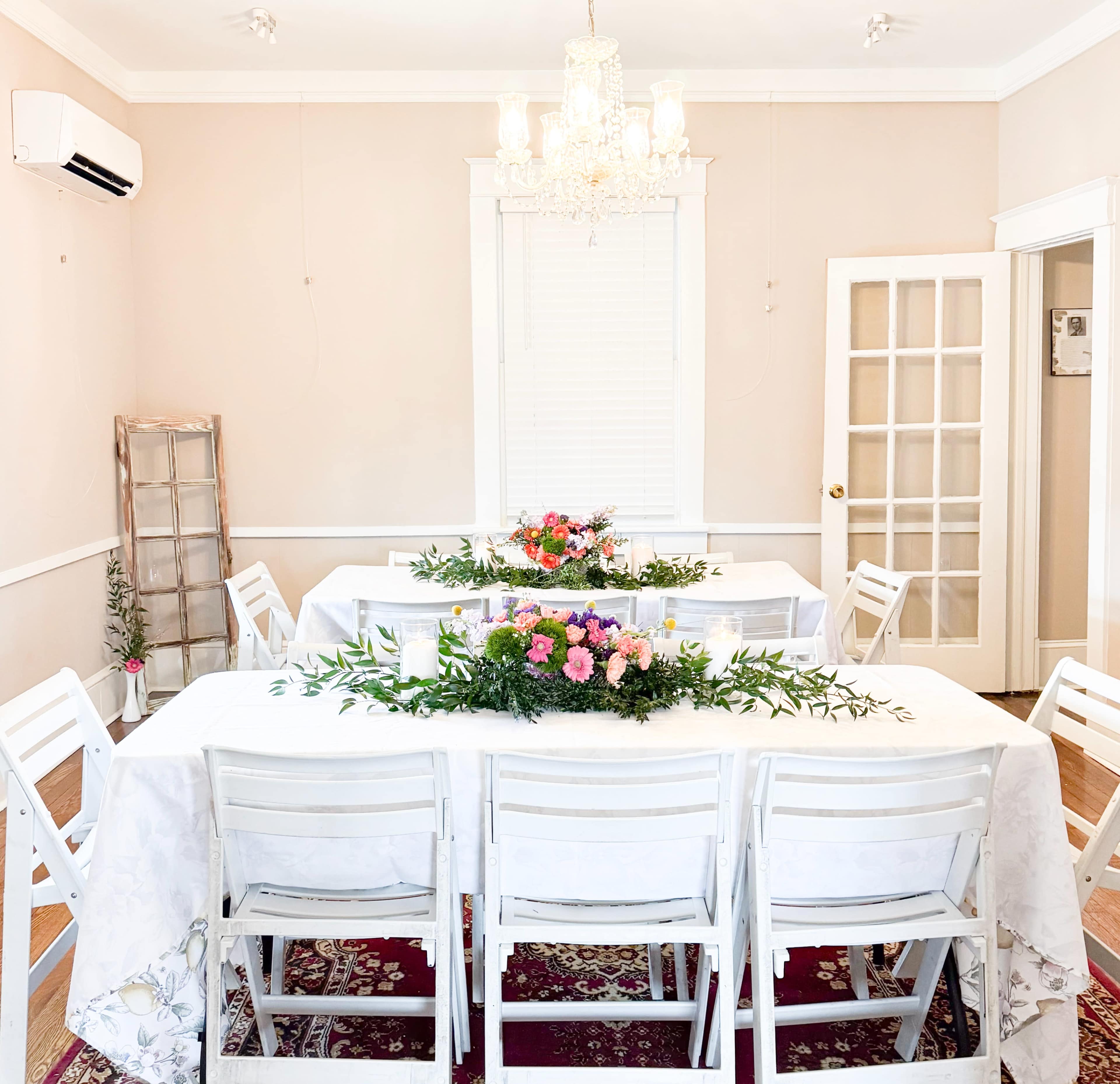 A dining room features two tables adorned with floral centerpieces and surrounded by white chairs under a chandelier.