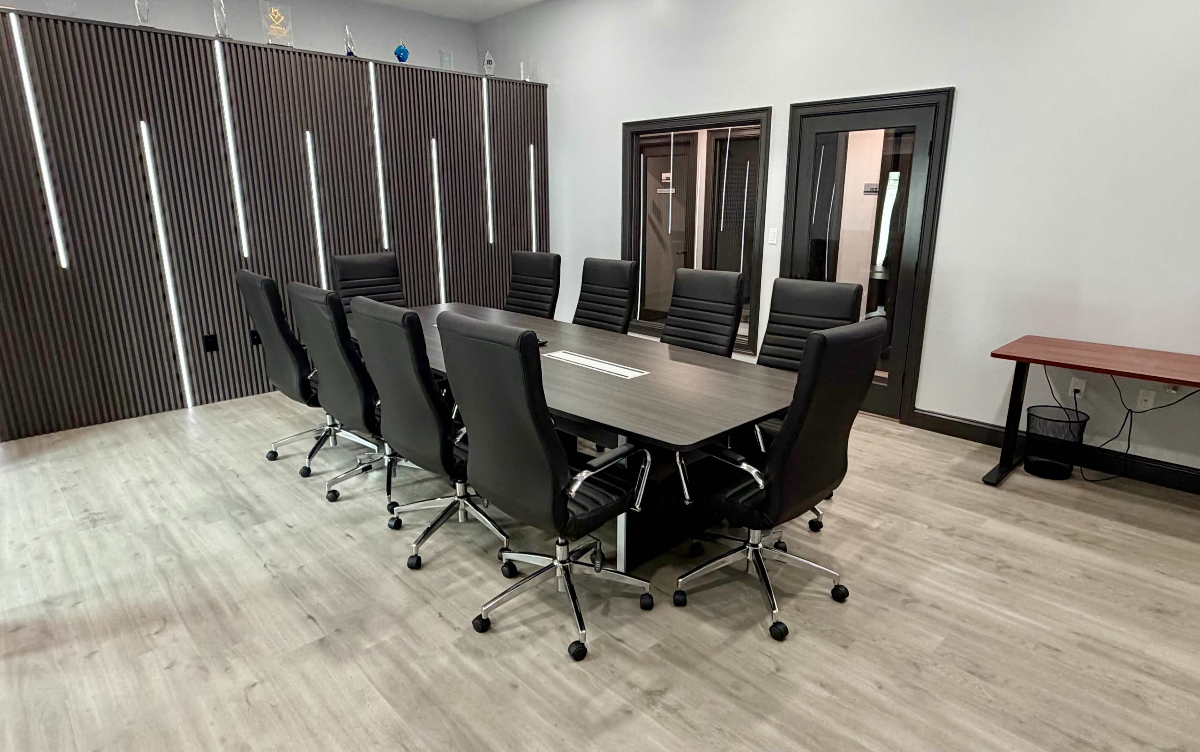 The image features a modern conference room with a large rectangular table surrounded by eight black leather chairs, complemented by wood-paneled walls and a simple wooden side table.