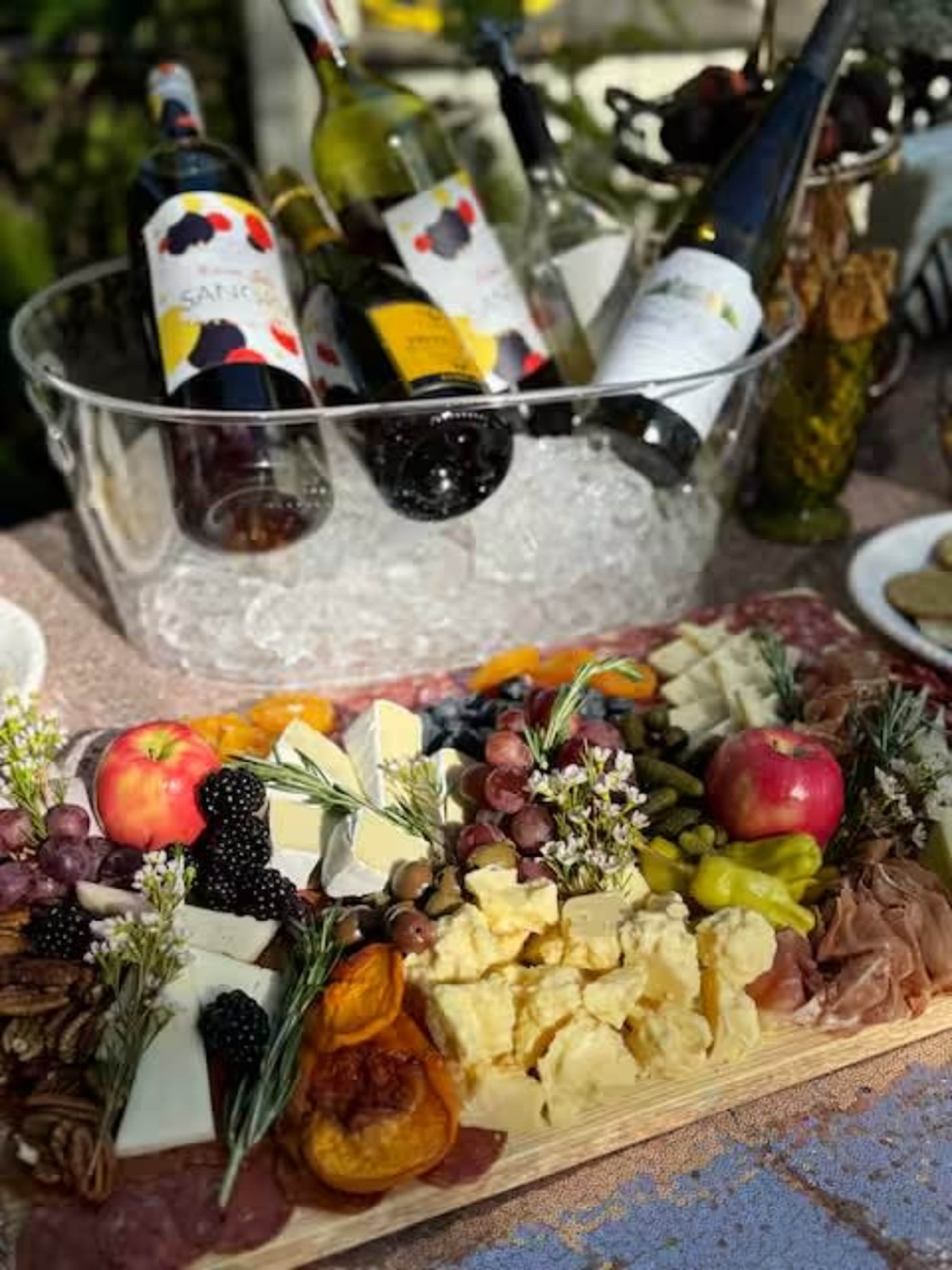 The image features a wooden platter filled with an assortment of cheeses, meats, fruits, and garnishes, alongside a bucket of wine bottles surrounded by ice.