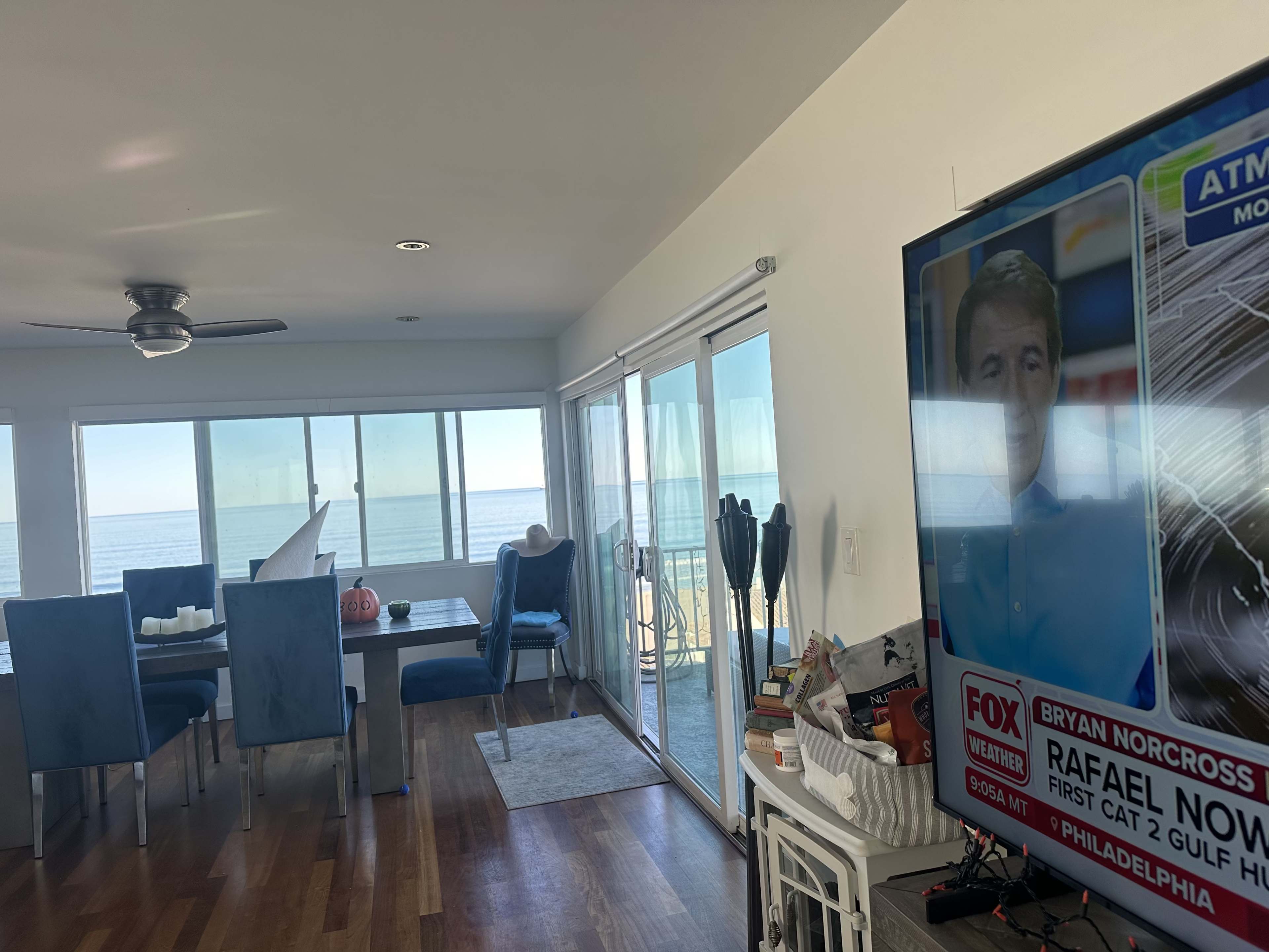 The image shows a modern interior of a beach house with a dining area, a large television displaying news, and a view of the ocean through large windows.