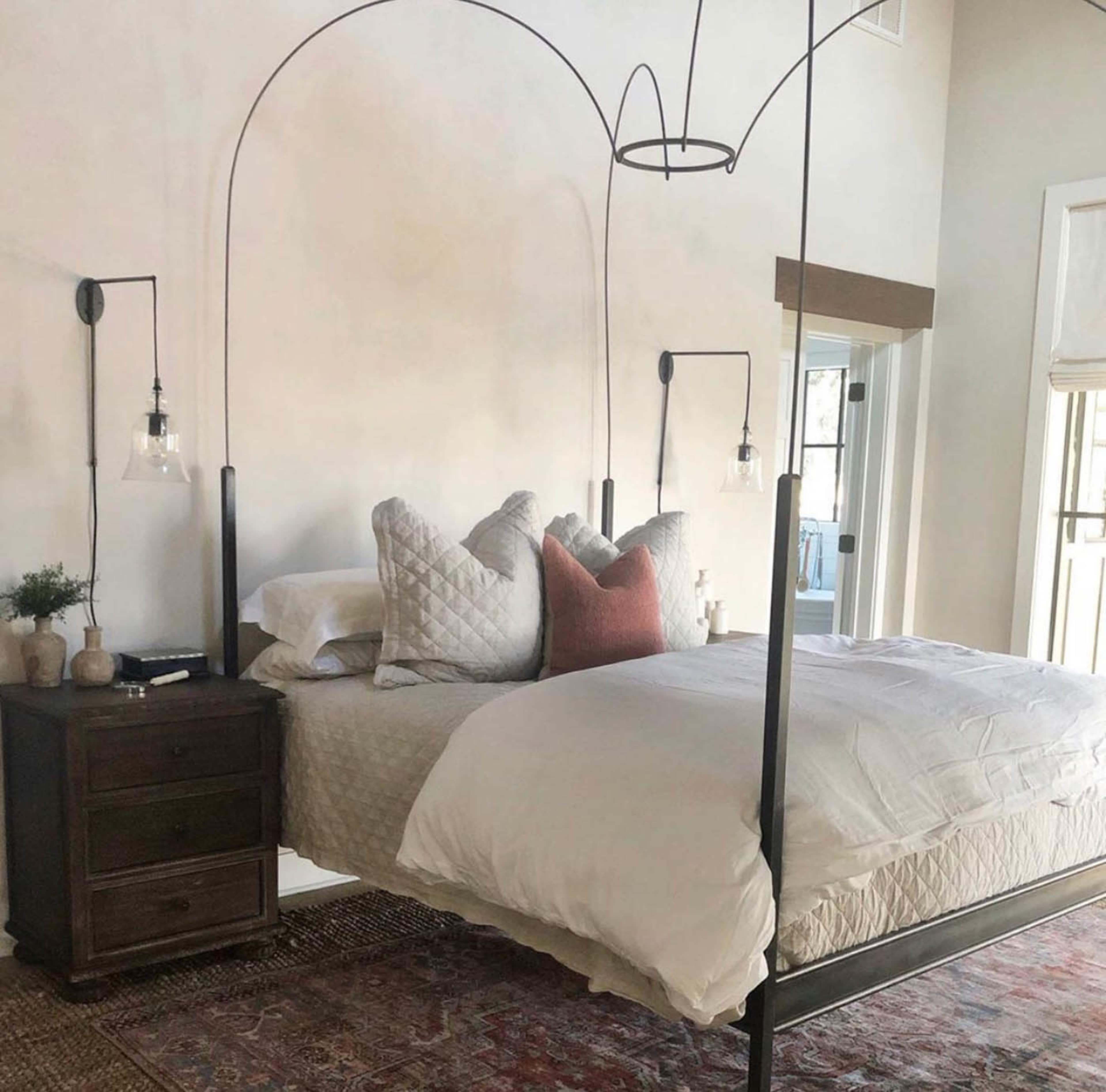 The image shows a modern bedroom featuring a metal canopy bed with a quilted white comforter and decorative pillows, accompanied by a bedside table and wall sconces.