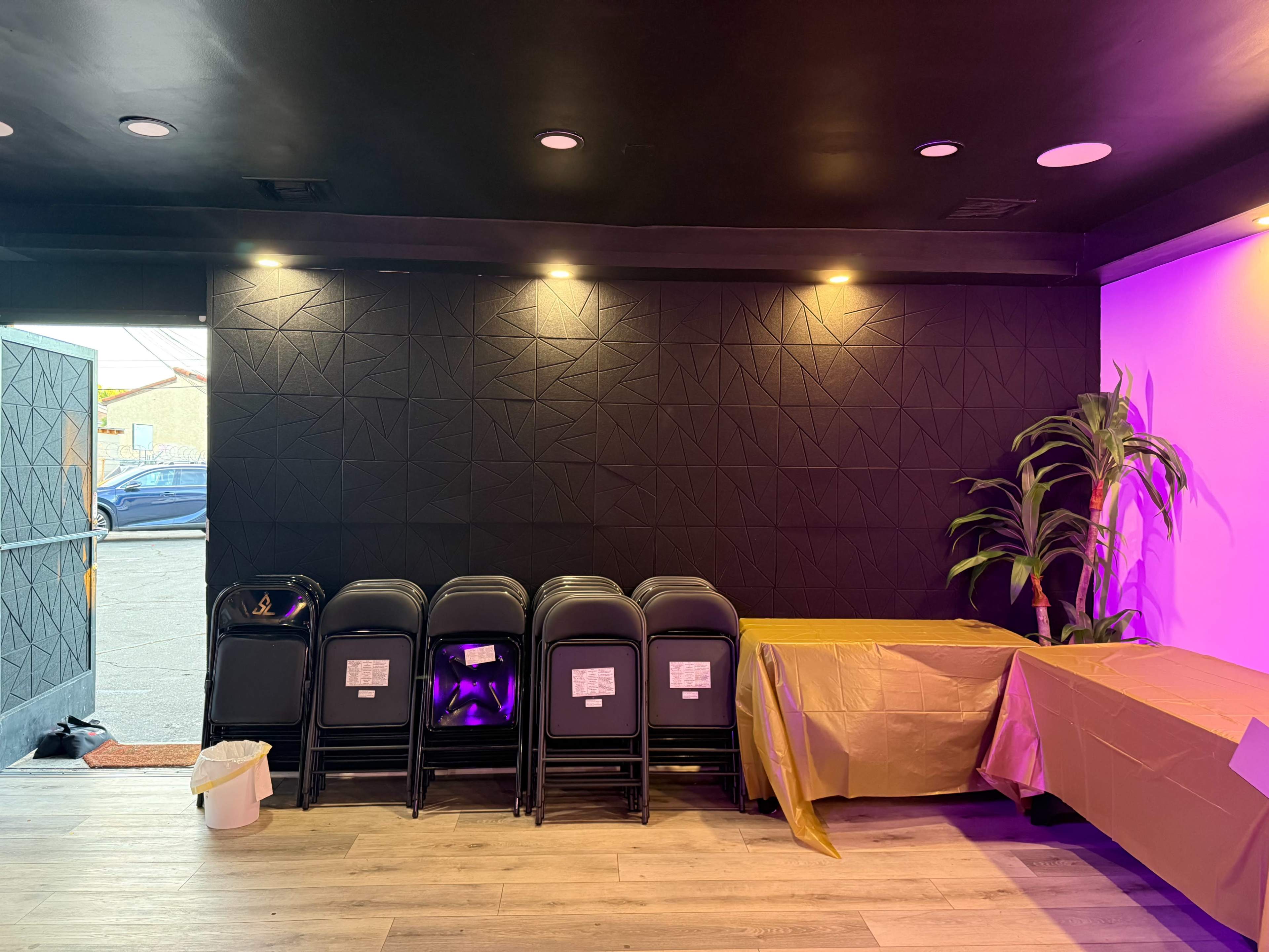 The image shows a room with a dark wall, several stacked black chairs, a table covered in a gold-colored cloth, and a green plant near an open door that reveals an outdoor space.
