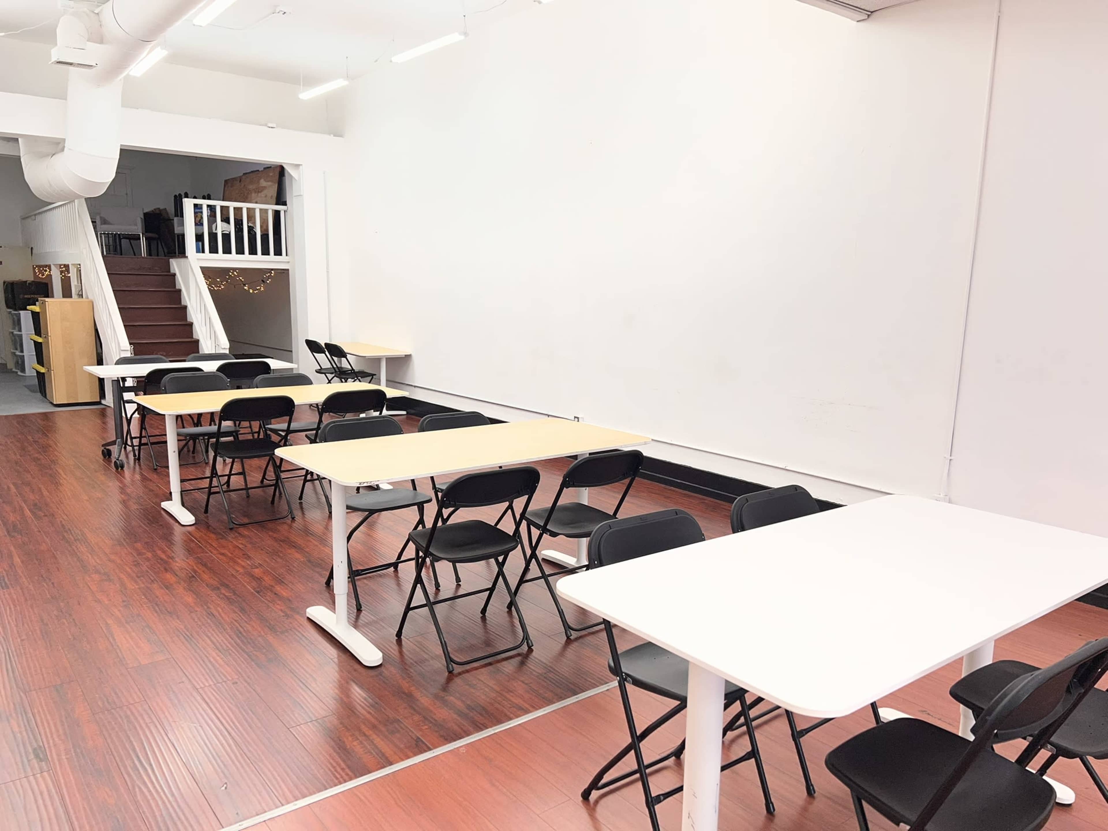 The image shows an empty room arranged with several tables and black chairs, along with a staircase in the background.