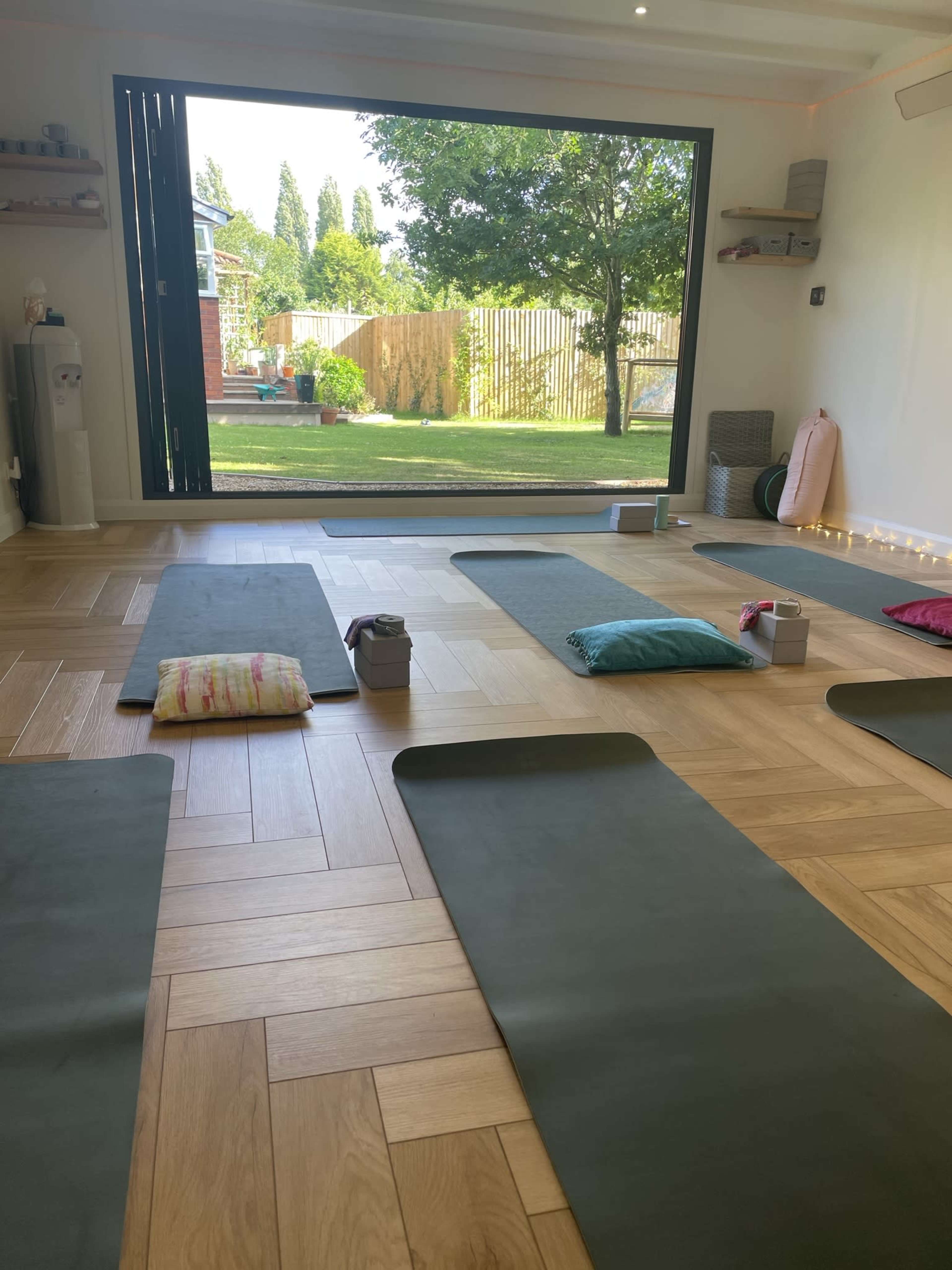 The image shows a yoga studio with green mats arranged on a wooden floor, overlooking a garden through a large window.